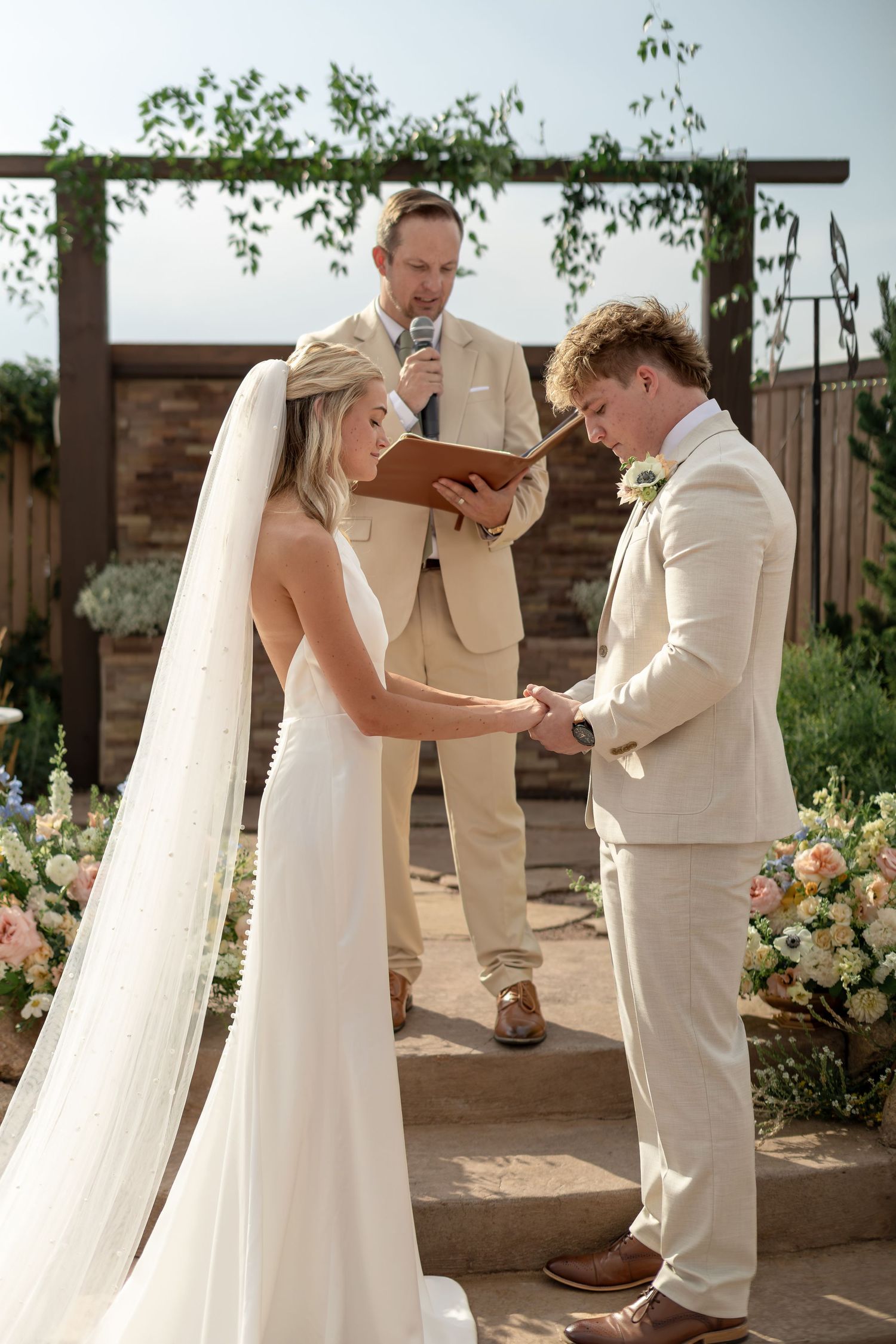 Bride and groom exchange vows at outdoor ceremony with officiant and floral arch backdrop.