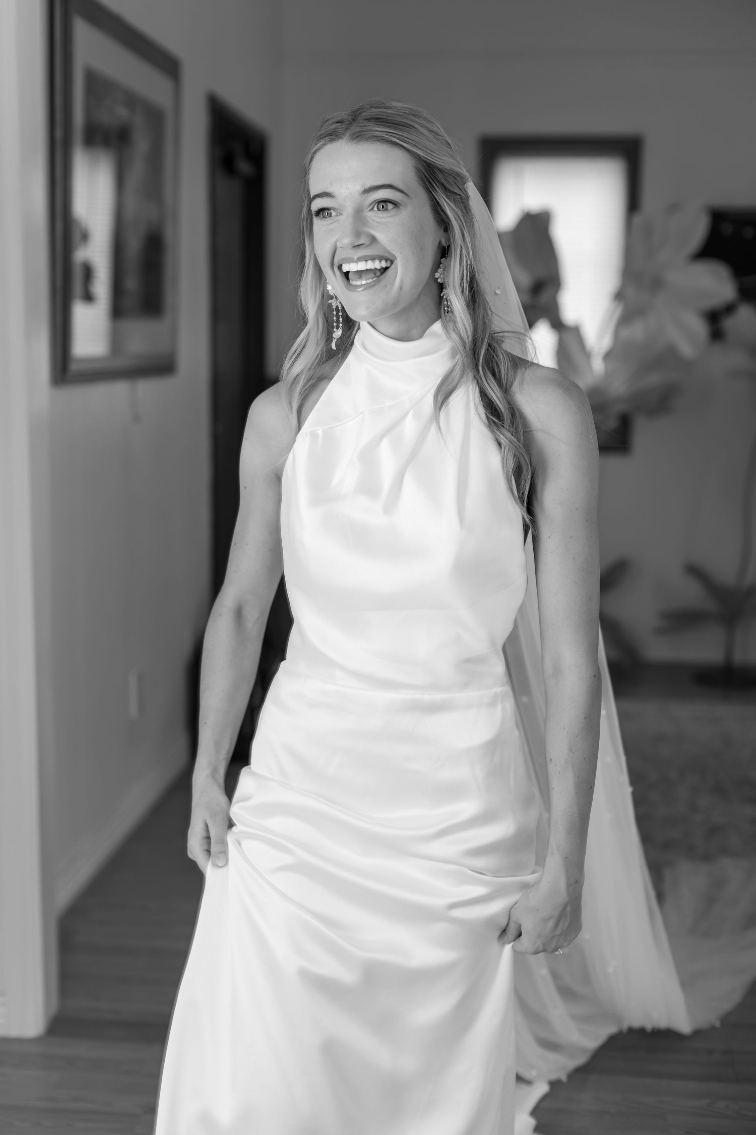 Black and white portrait of radiant bride laughing in white halter wedding gown indoors.