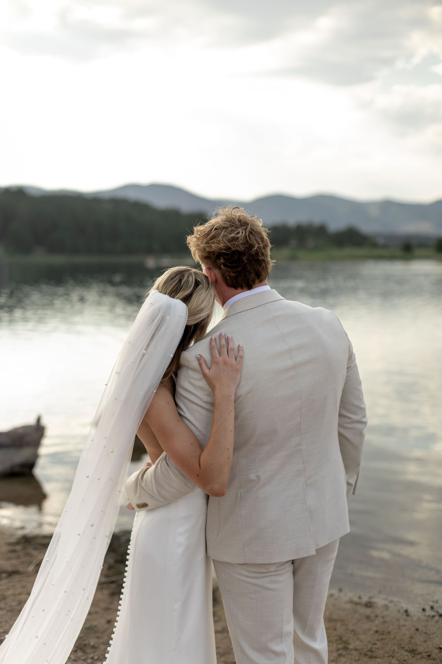 Bride and groom embrace while gazing at a serene mountain lake at sunset on their wedding day.