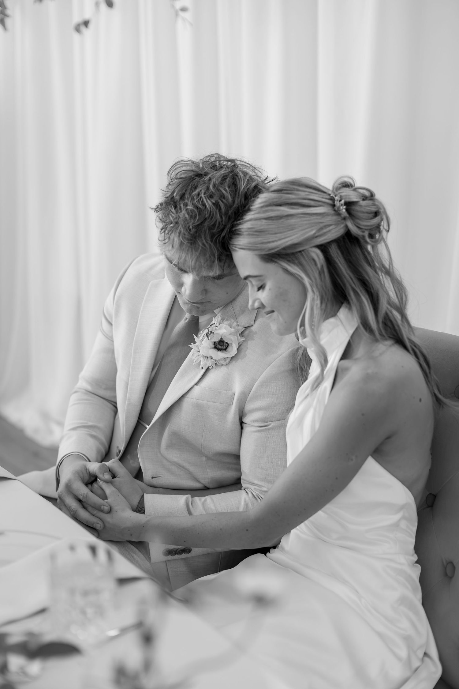 Black and white photo of bride and groom sitting closely together holding hands at reception table.