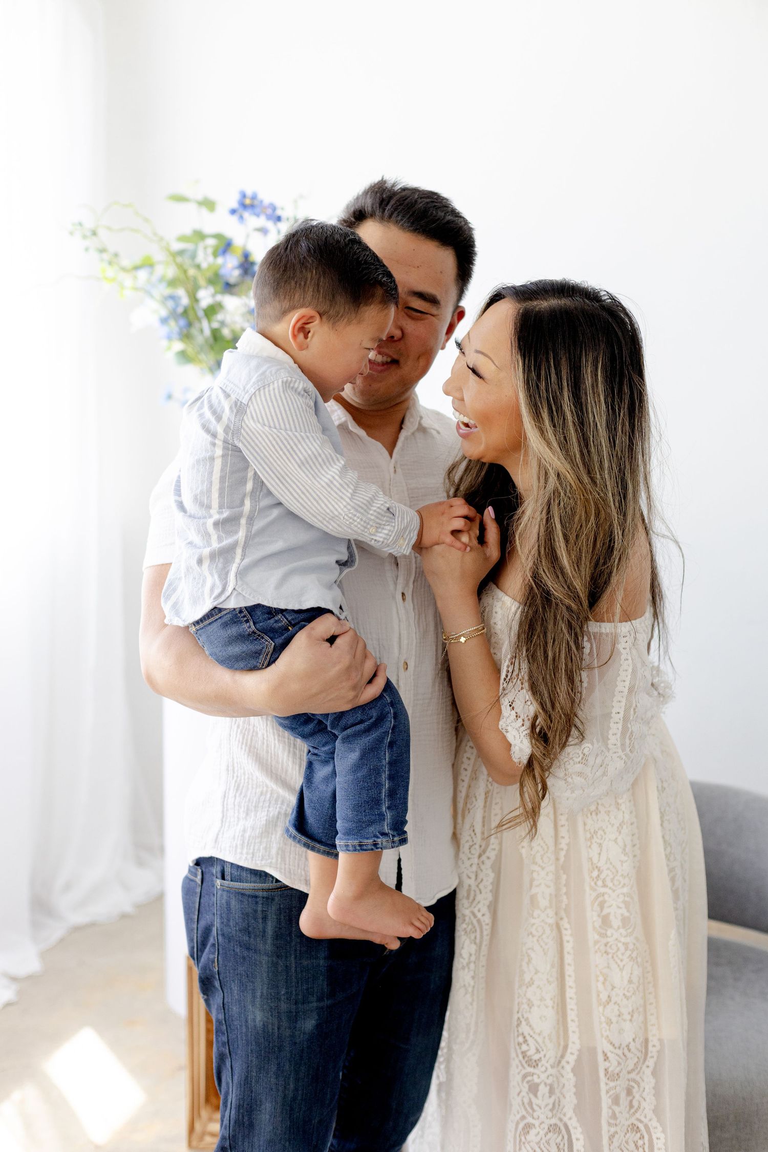 A happy Asian family with a father holding their young son while smiling with the mother in a bright white room.