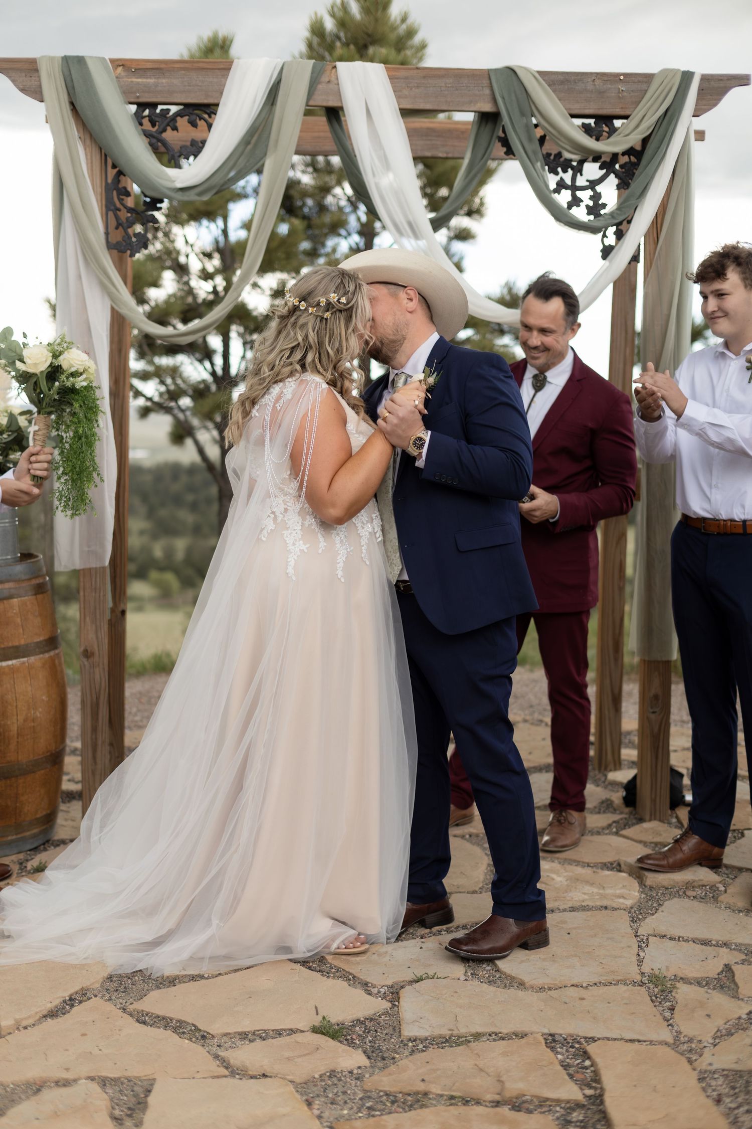 Bride and groom share first kiss under rustic wooden arch with sage green draping at outdoor wedding ceremony.