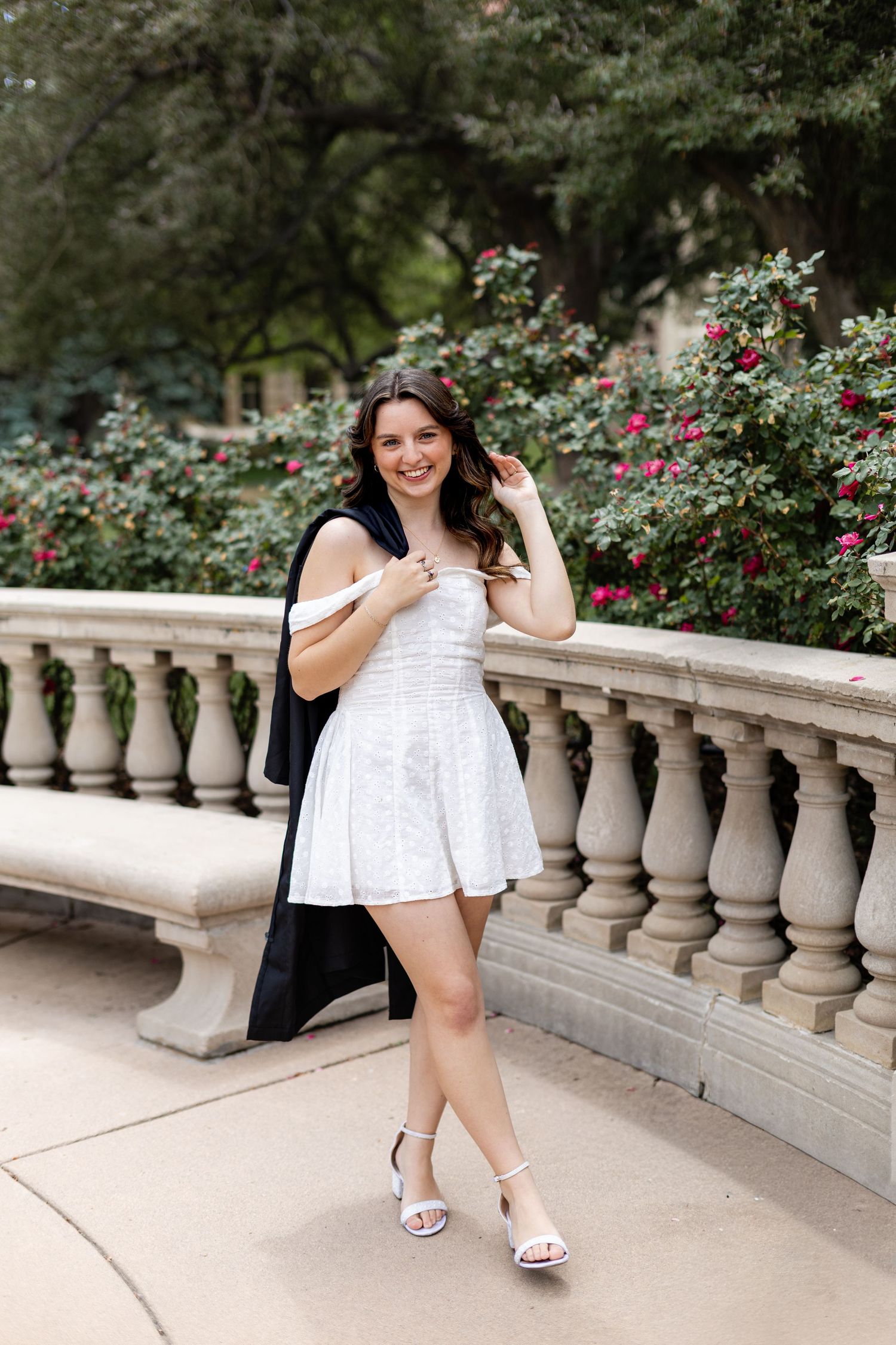 Young woman in white off-shoulder mini dress and heels posing by stone balustrade with red roses.