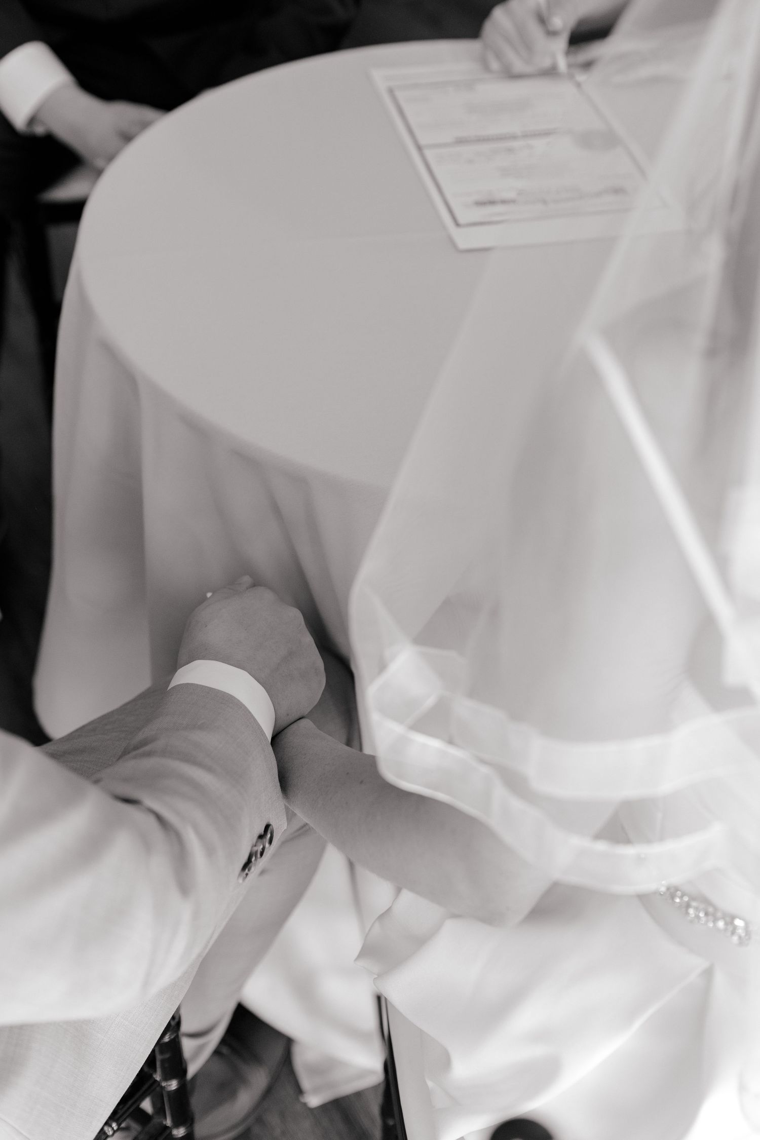 Black and white photo of bride and groom holding hands while signing wedding certificate at a draped table.