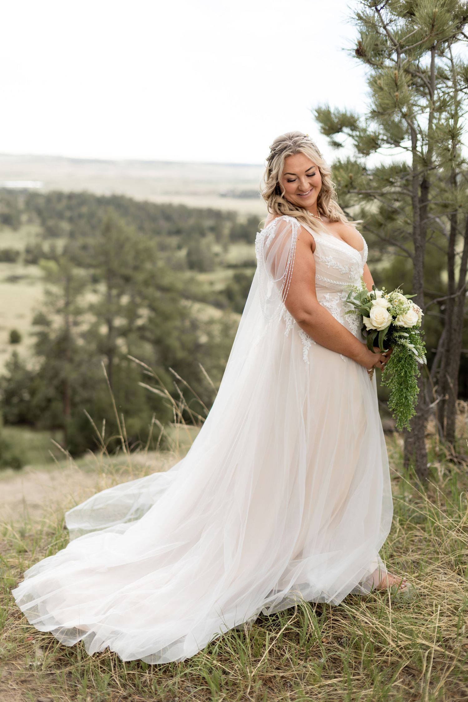 Bride in flowing white tulle gown holds bouquet on scenic hilltop surrounded by pine trees.