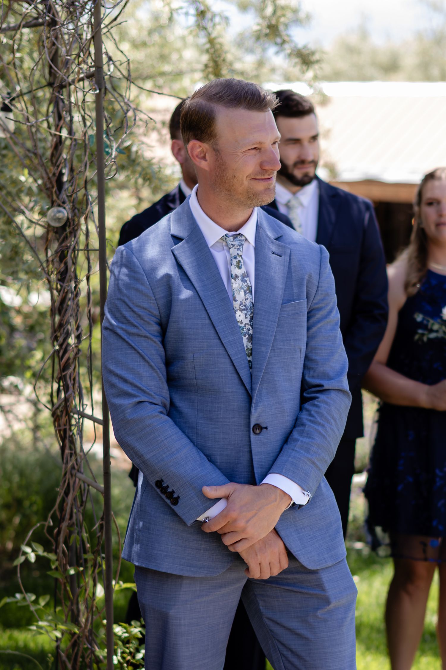Groom in light blue suit with floral tie smiling at outdoor wedding ceremony with guests in background.