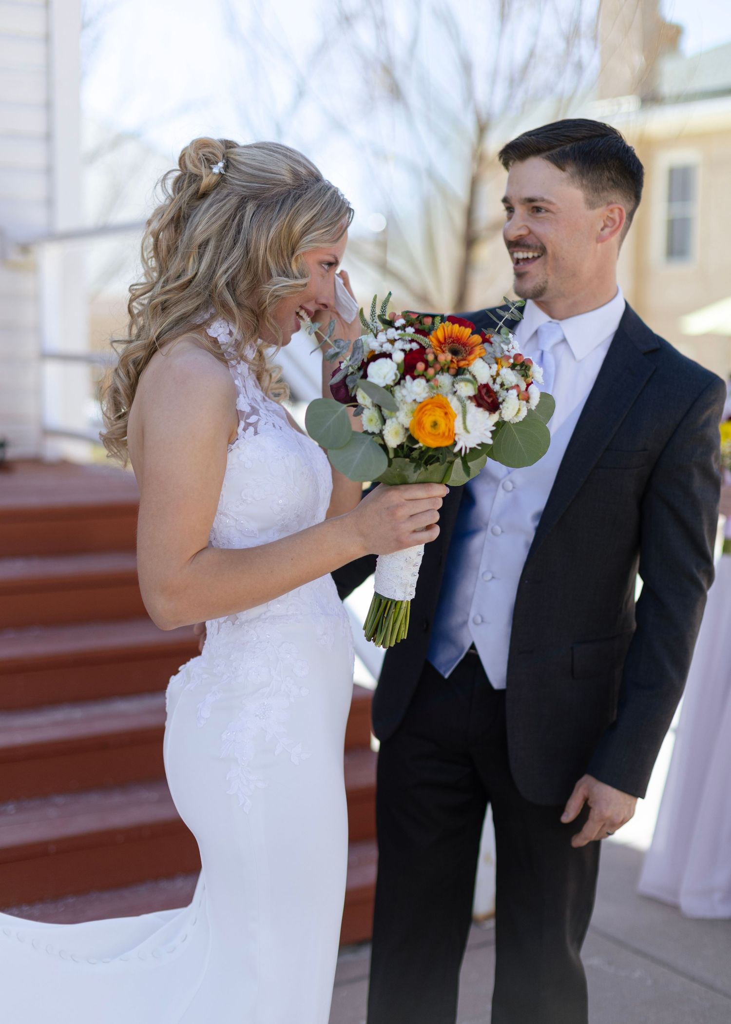 Smiling bride in lace gown and groom in navy suit sharing joyful moment with bouquet outdoors.