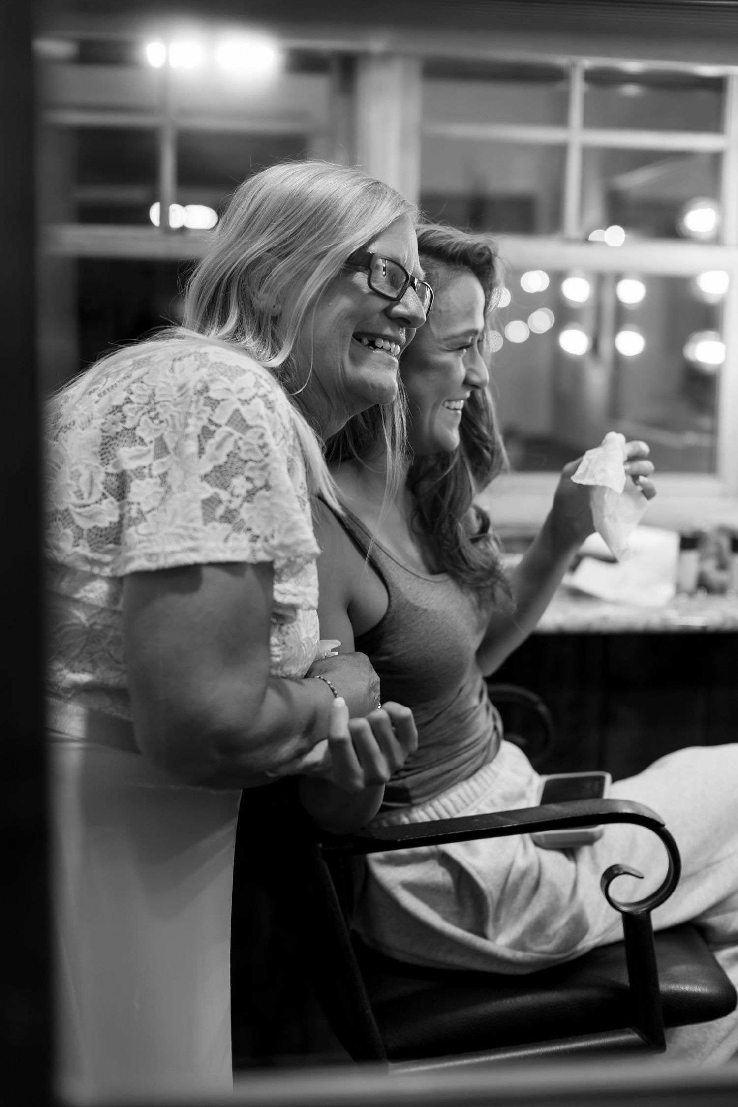 Two women smile and laugh together at an indoor wedding reception in black and white photography.