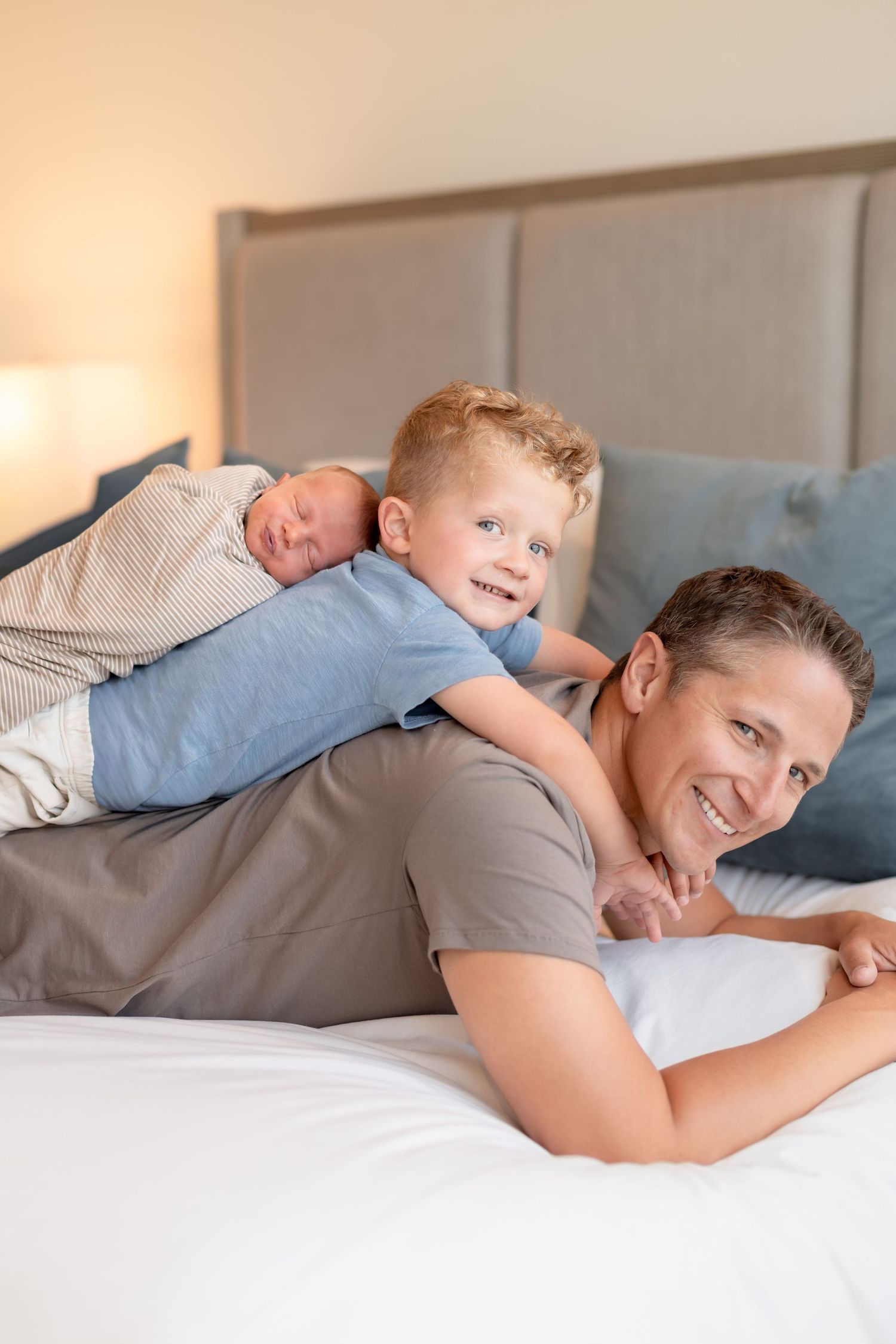 A smiling father lies on a bed with his young redhead son and swaddled newborn baby in a cozy bedroom.
