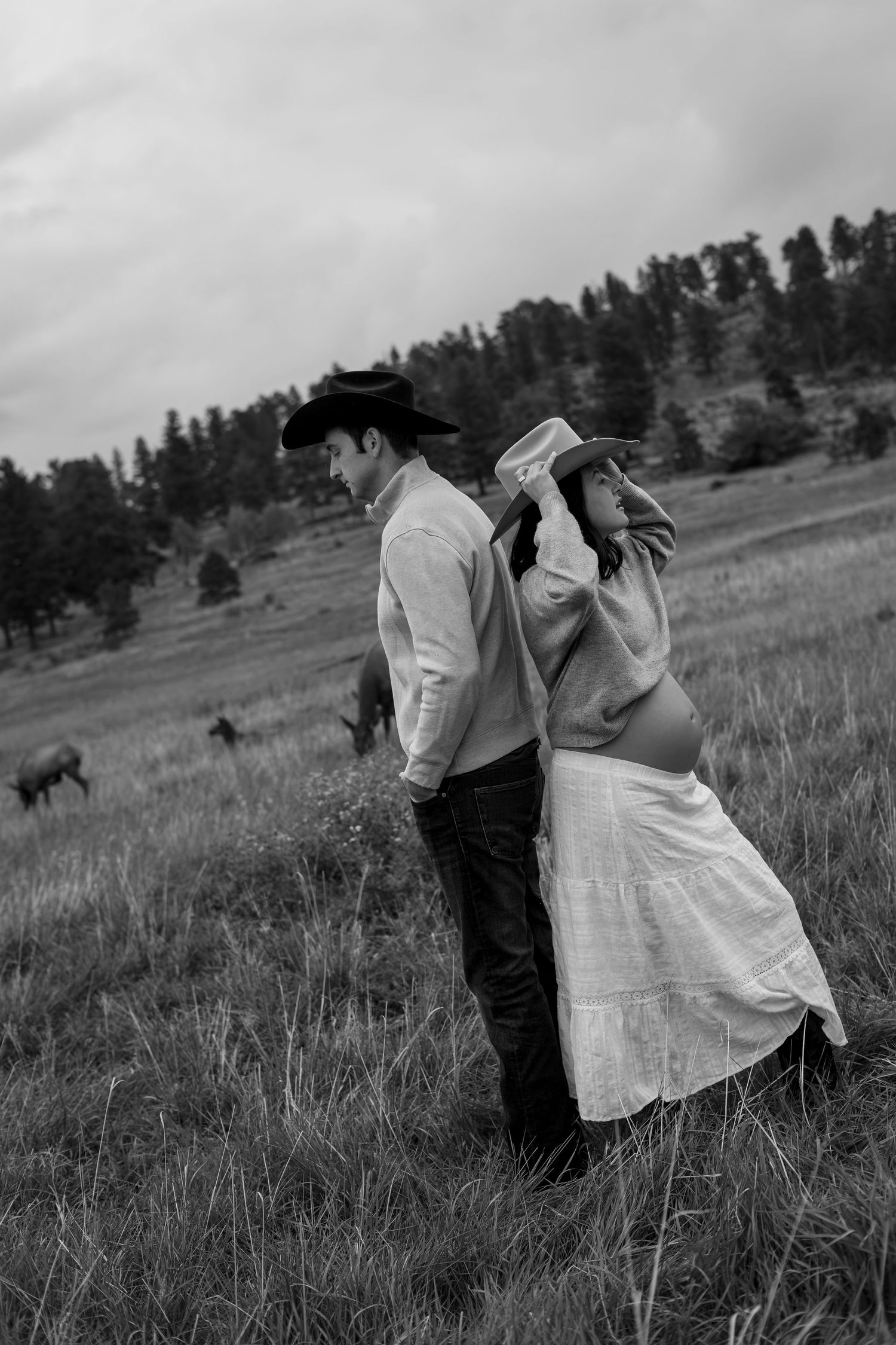 Black and white photo of a pregnant couple in cowboy hats standing in a field with horses and trees in background.