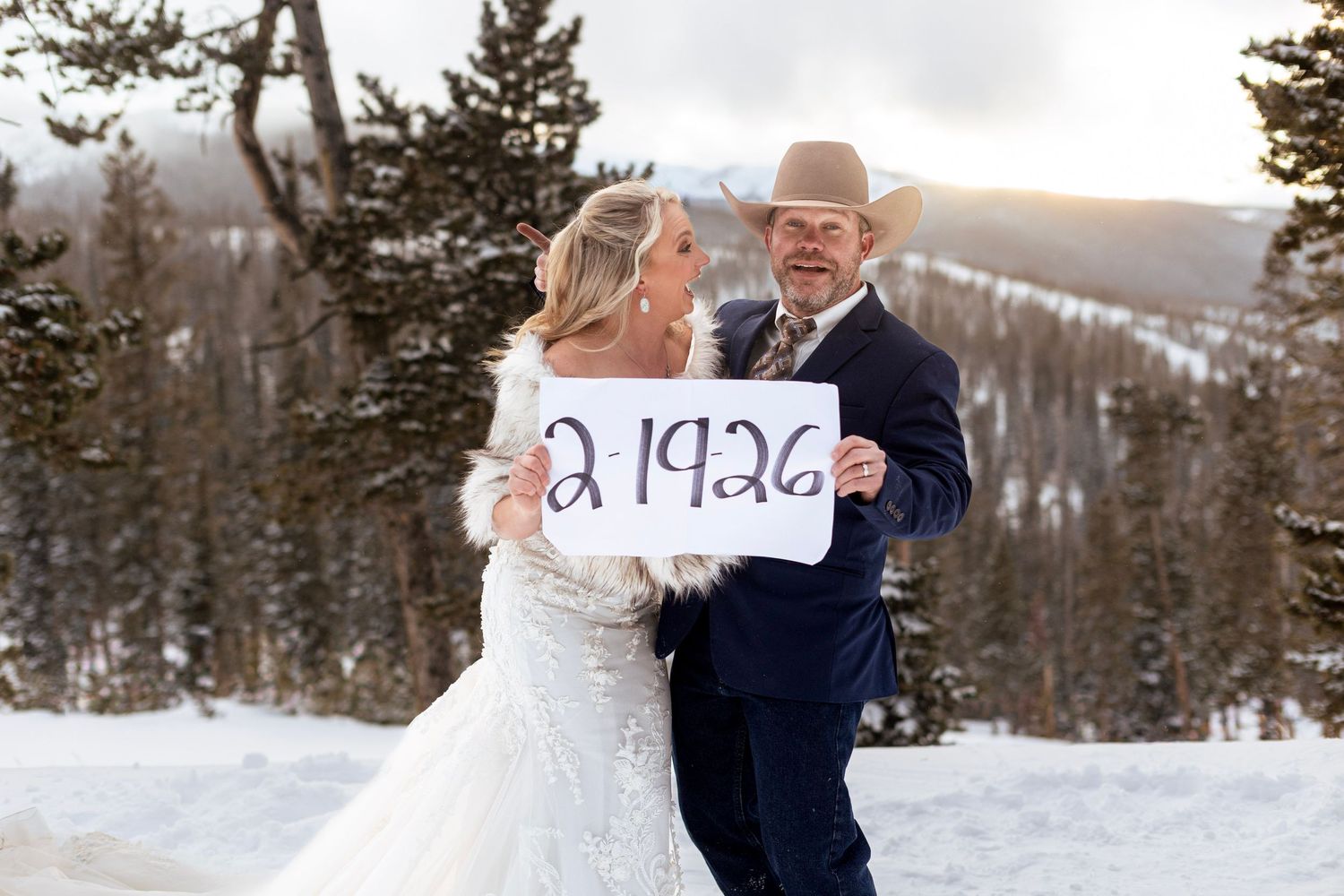 A couple in wedding attire shares a romantic moment in a snowy winter landscape with evergreen trees in the background.