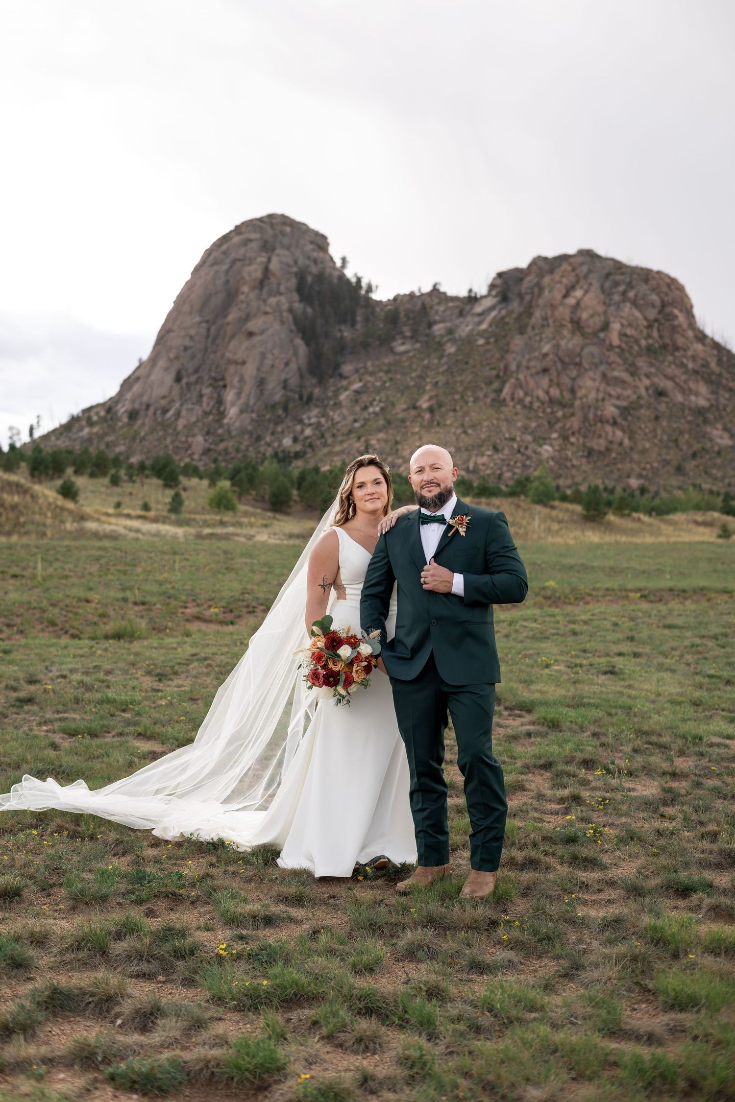 Bride and groom pose together in a mountain meadow, she in a white gown with veil, he in a dark green tuxedo.