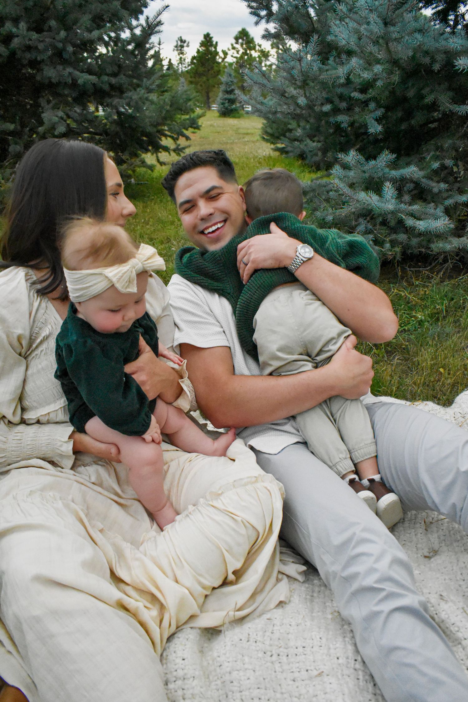 A family cuddles together on a white blanket with evergreen trees in the background.