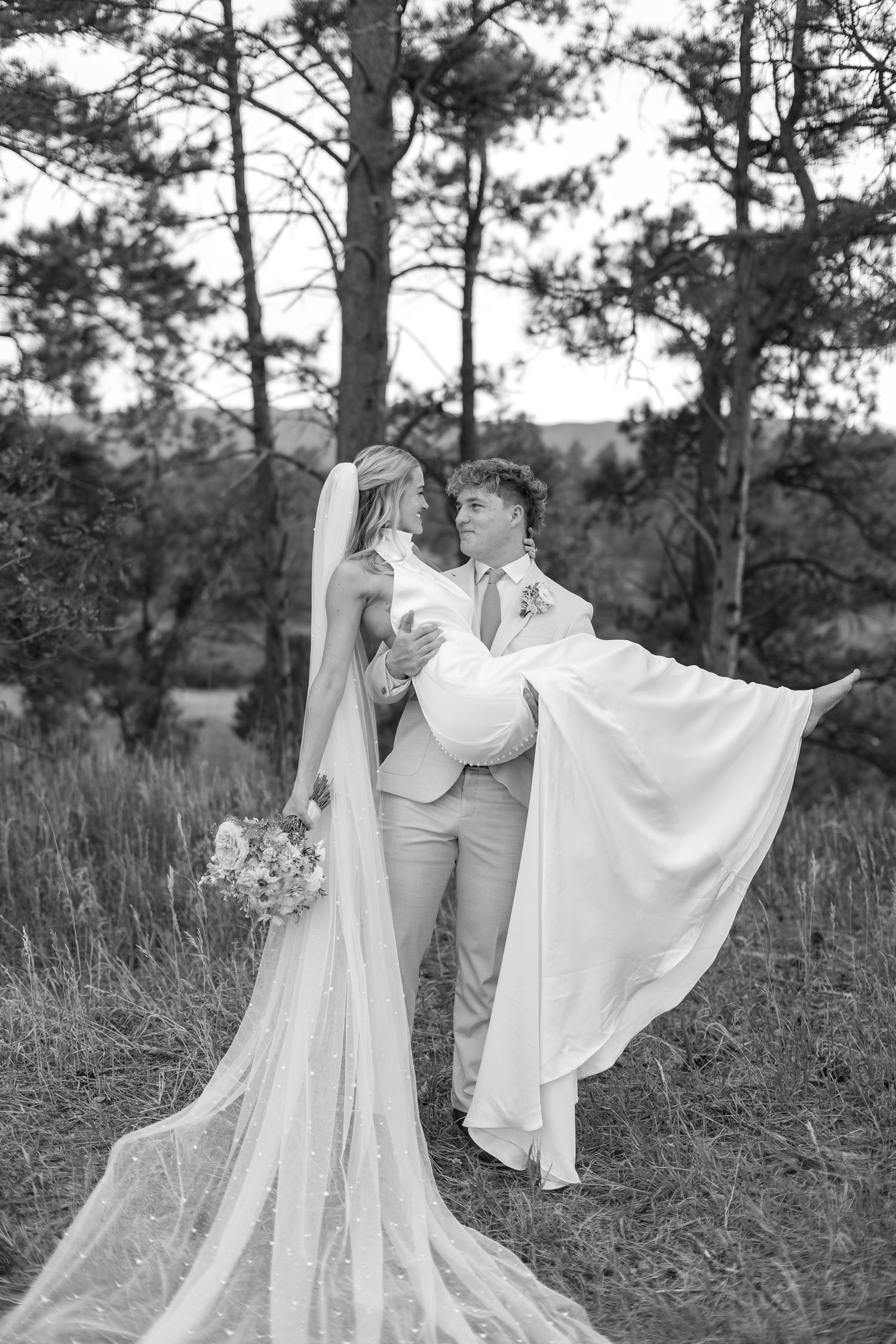 Black and white photo of groom carrying bride in flowing wedding dress and veil among tall pine trees.