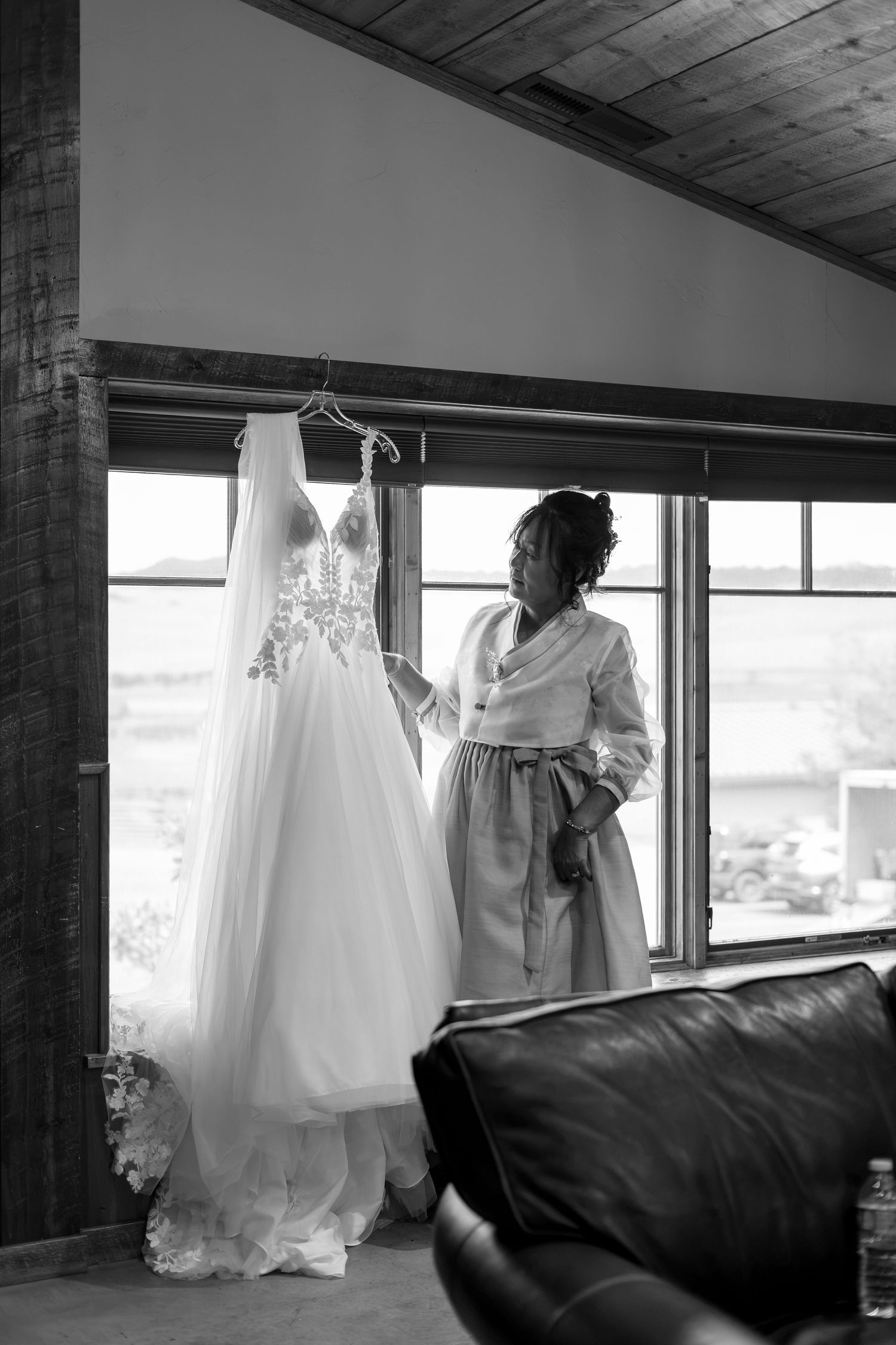 Bride admiring her elegant lace wedding gown hanging by a window in black and white photo.