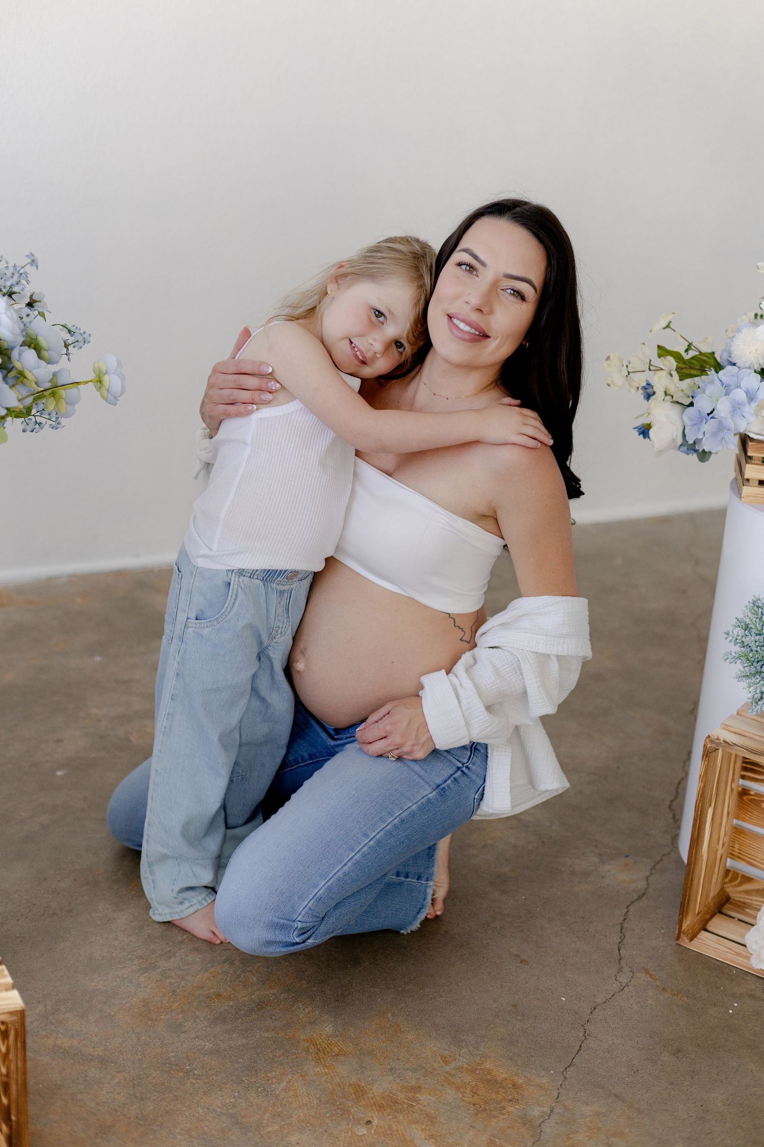 Pregnant woman in white crop top and jeans kneeling with young blonde girl hugging her, surrounded by blue flowers.