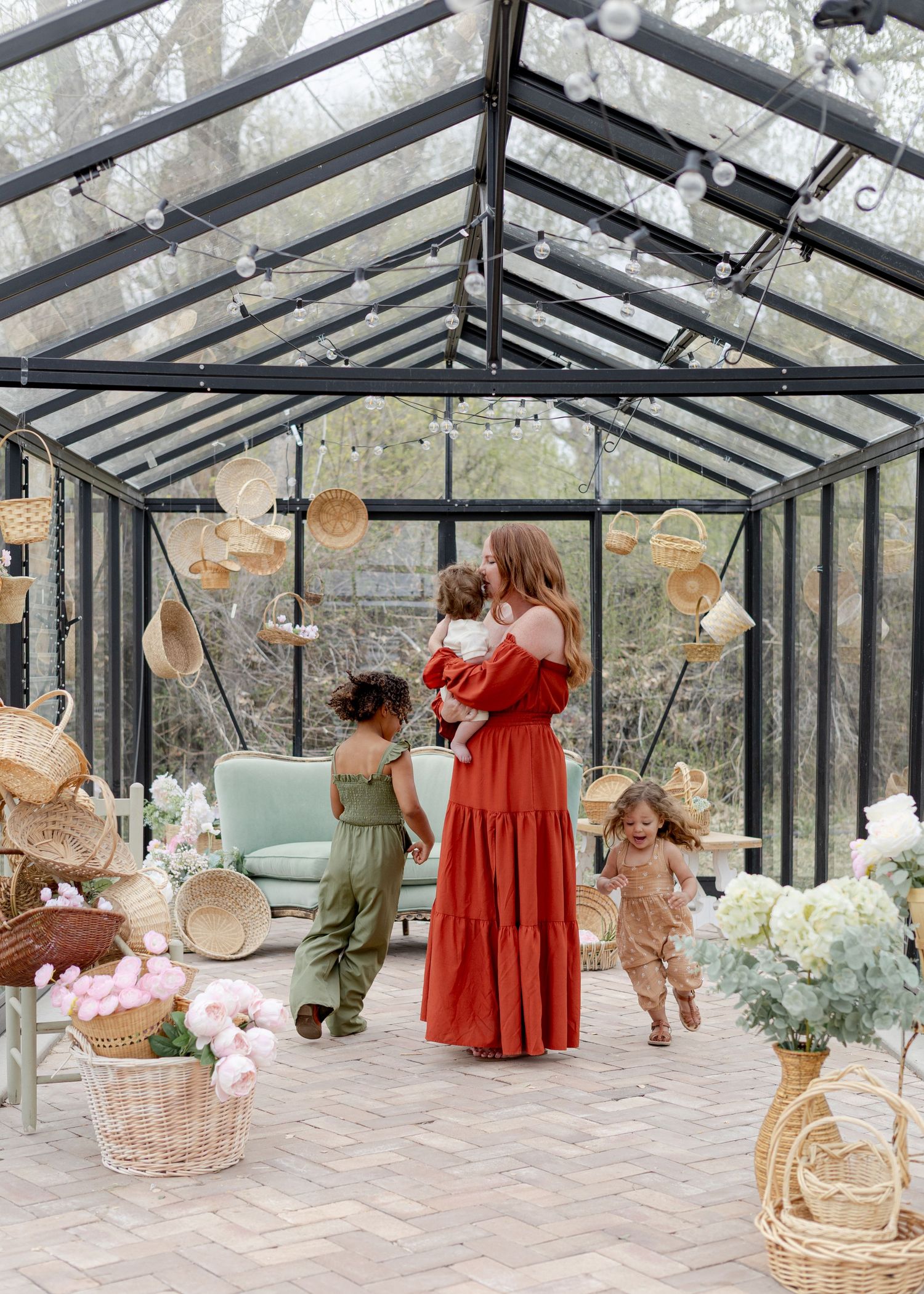 Woman in red dress holds child in glass greenhouse with wicker decor, flowers, and two toddlers nearby.