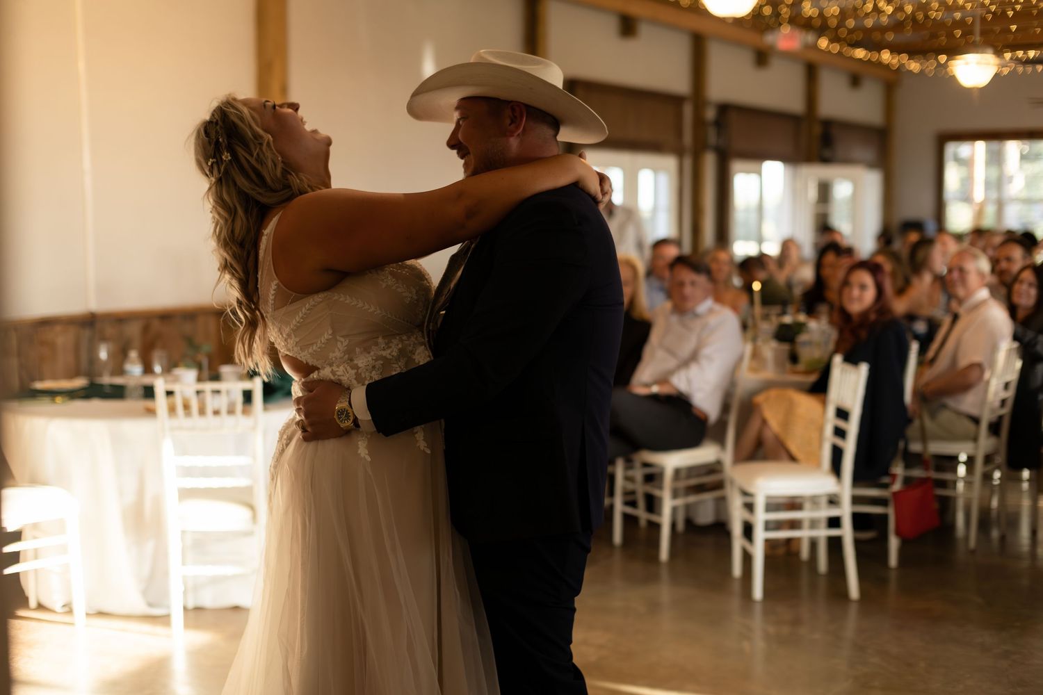Bride and groom share their first dance in a warmly lit wedding reception hall with guests watching.