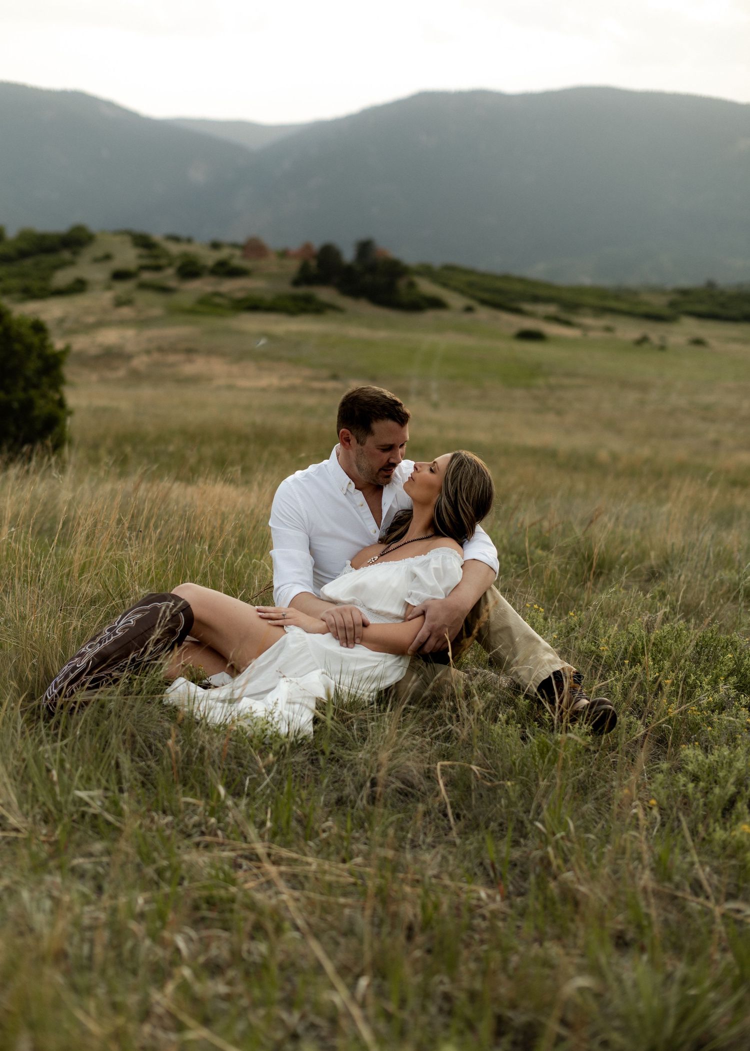 Couple sitting in a lush green meadow with mountain backdrop, sharing a romantic moment during an outdoor engagement session.