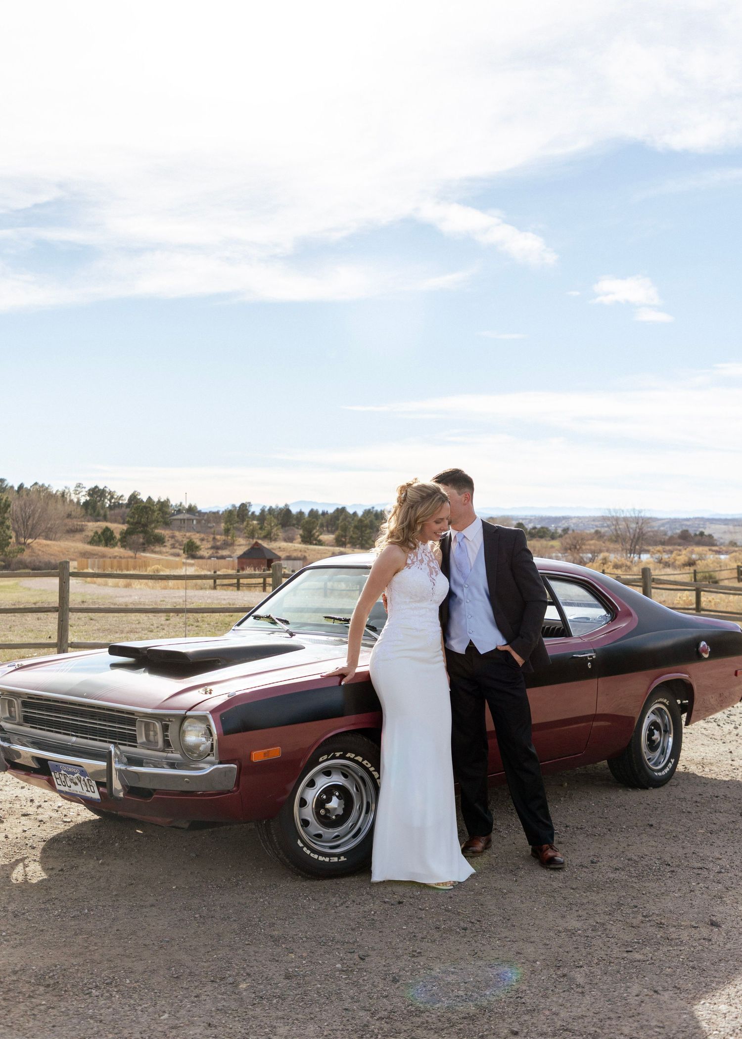 A bride and groom share a romantic moment leaning against a classic red muscle car outdoors.