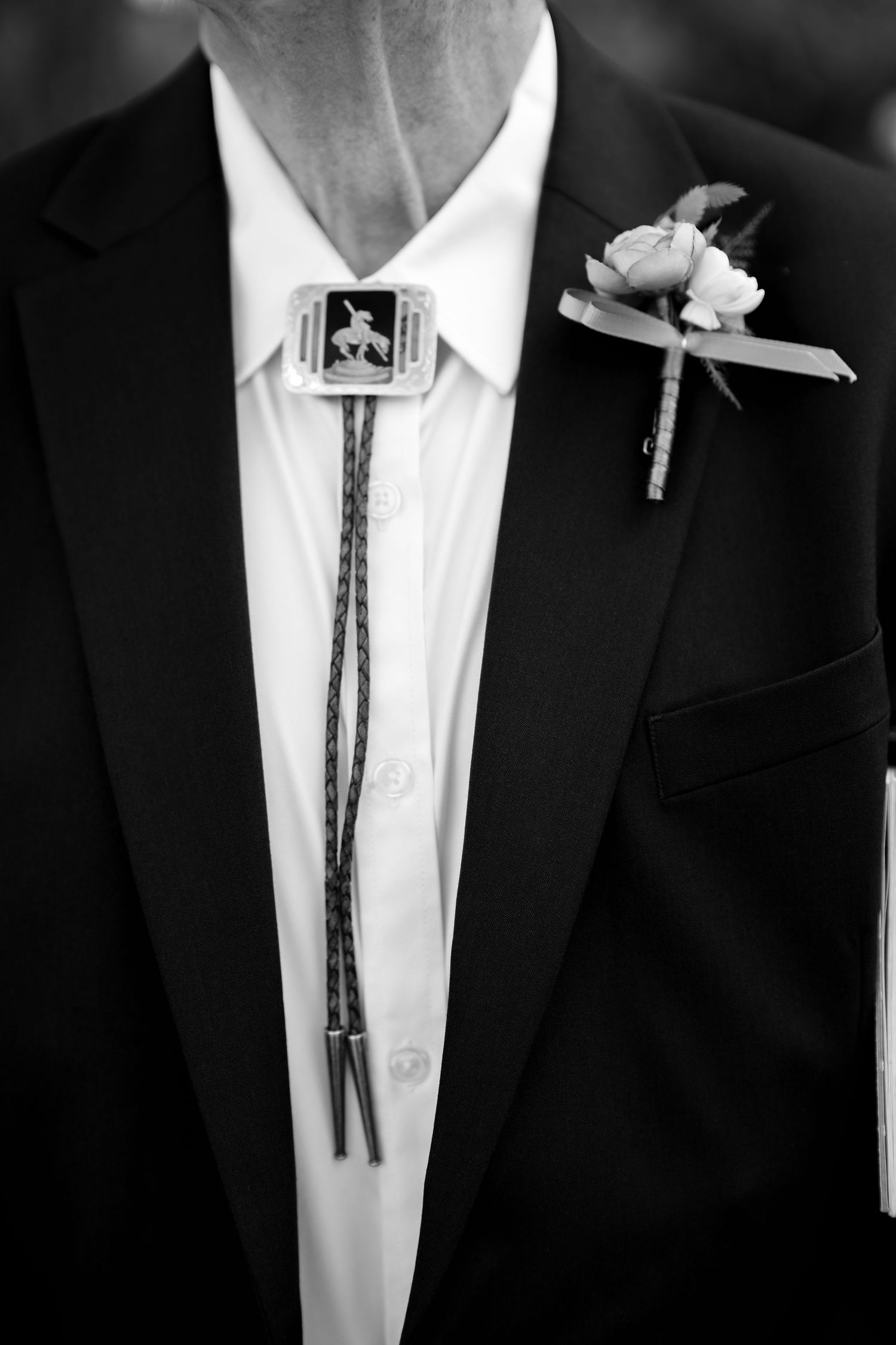 Groom wearing a bolo tie with western buckle and floral boutonniere on dark suit jacket.