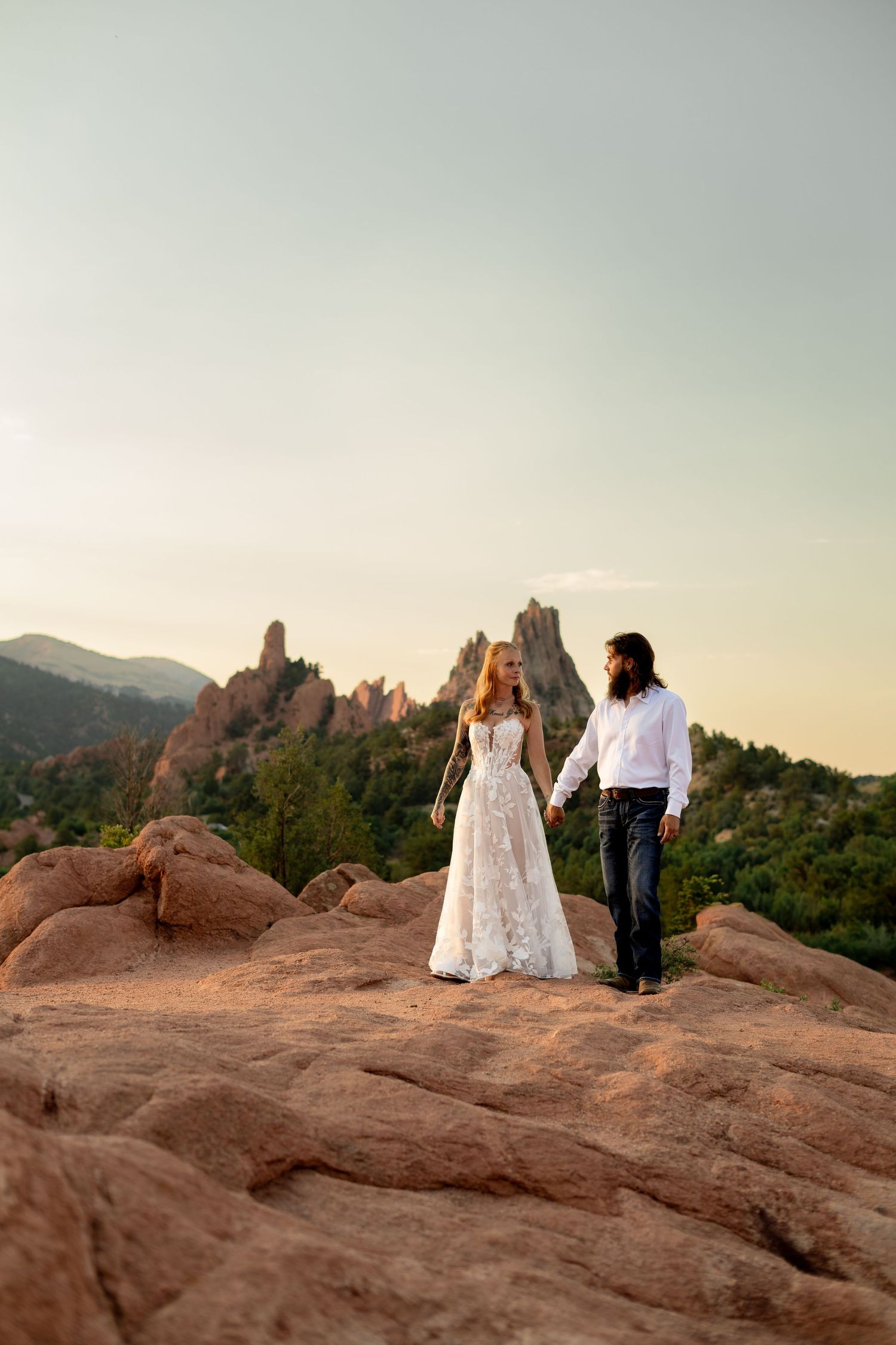 Bride and groom hold hands on red rocks at Garden of the Gods, Colorado, with dramatic rock formations at sunset.