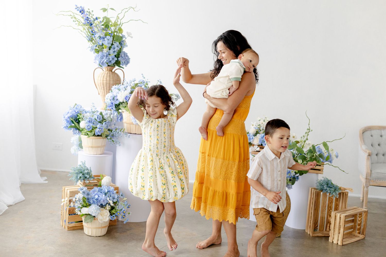 A mother in a yellow dress dances with her three children surrounded by blue hydrangeas in a bright white studio.