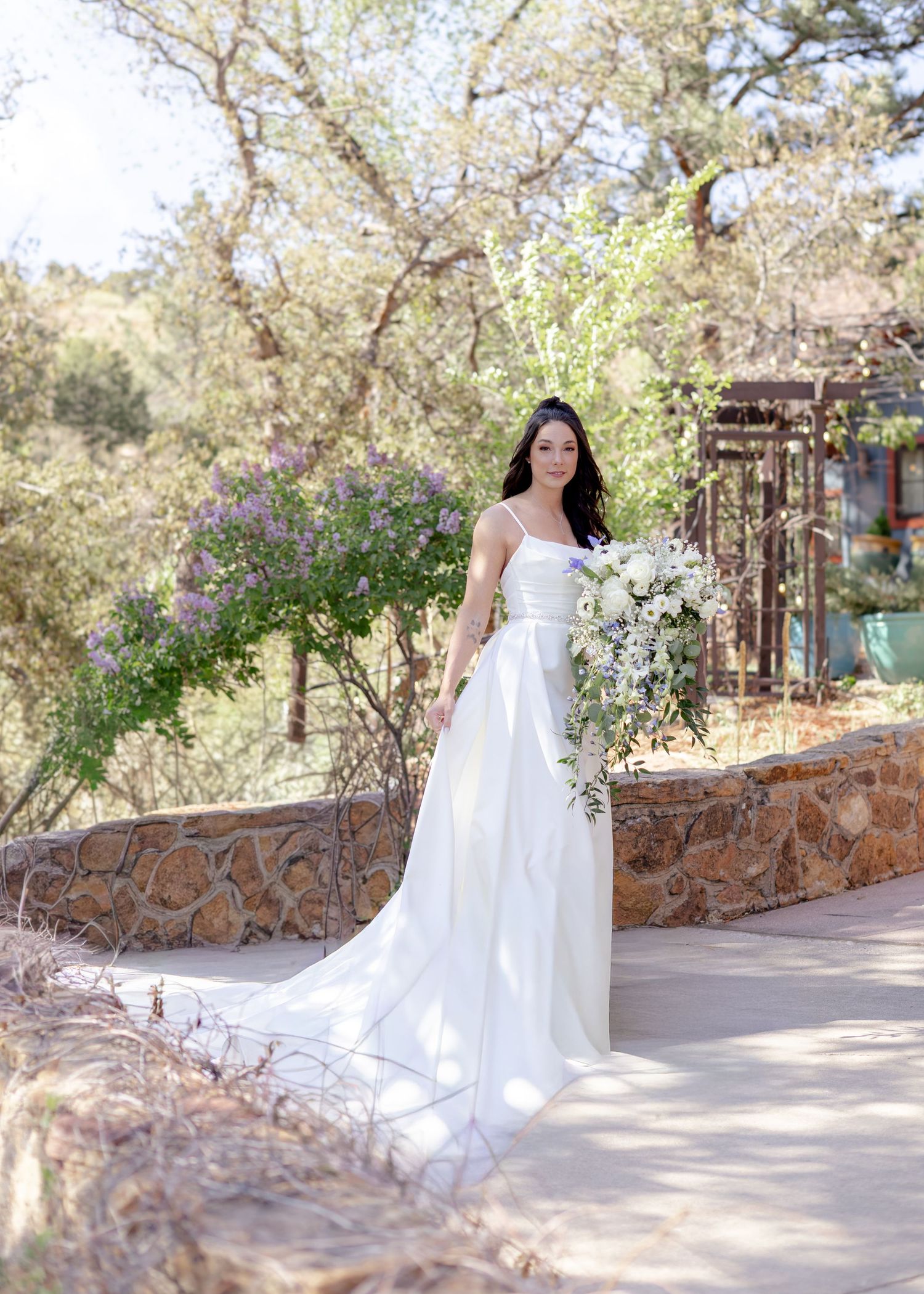 Bride in white spaghetti strap gown holding floral bouquet, posing outdoors among trees and stone wall.