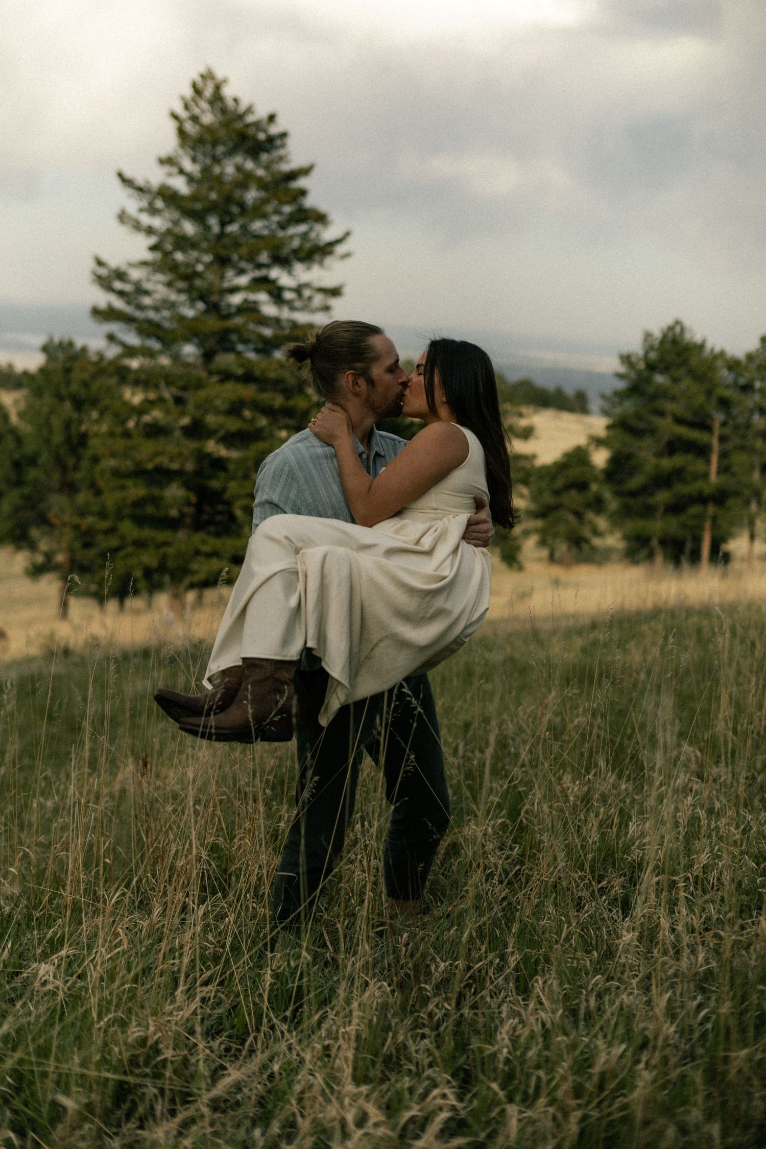 Man carrying woman in white dress sharing a kiss in a lush green meadow with pine trees.