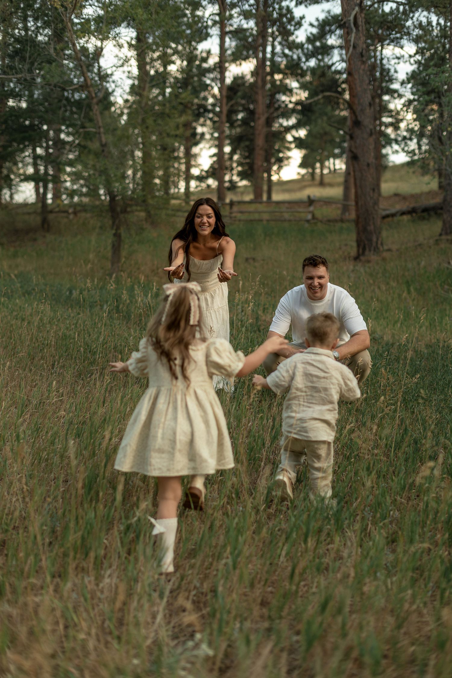Family playing in a green meadow surrounded by pine trees, parents welcoming children running toward them at sunset.