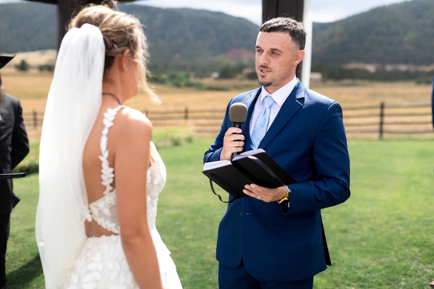 Groom in a navy suit reads vows to his bride outdoors at a rustic wedding ceremony.