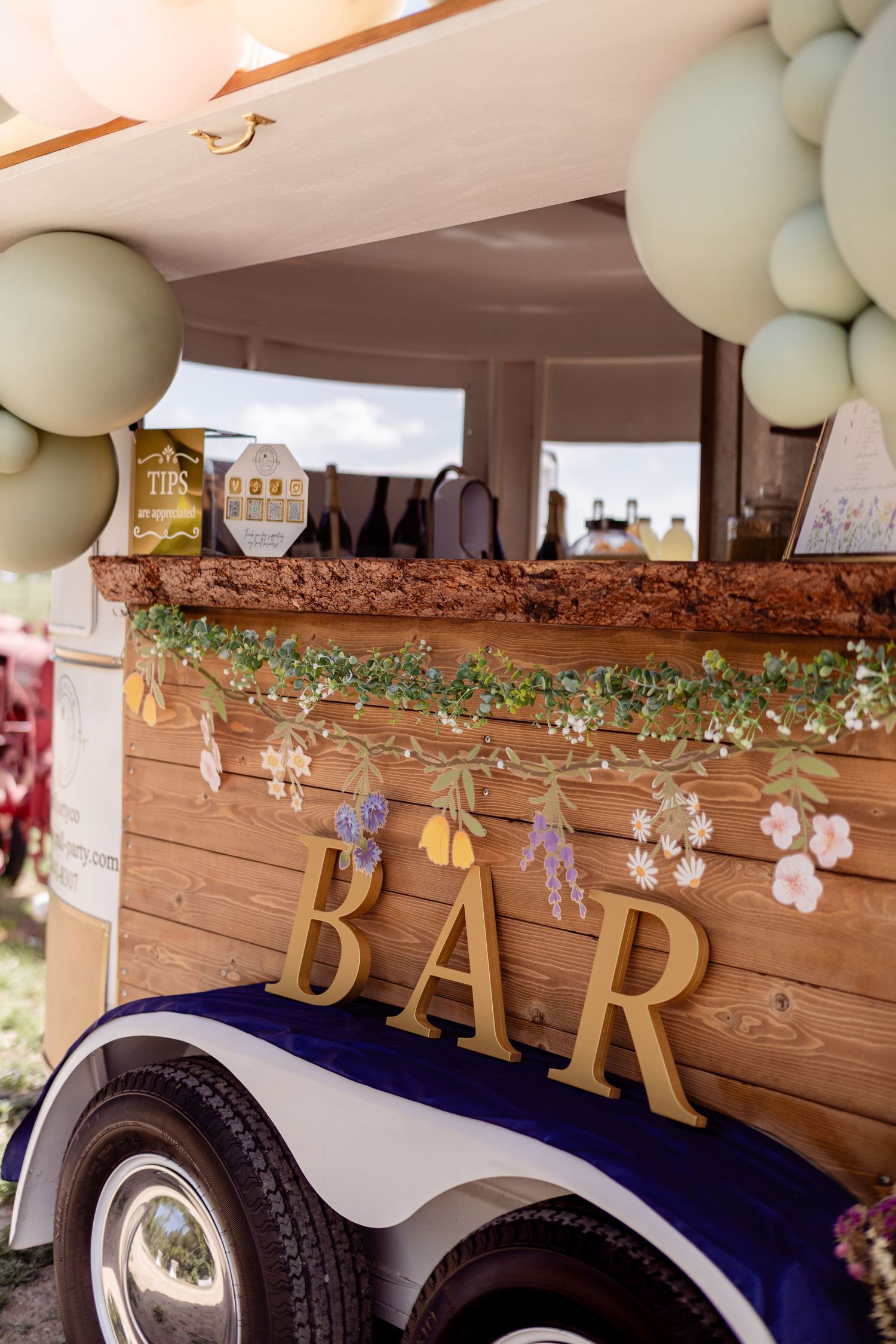 Rustic wooden bar cart decorated with flowers and balloons at an outdoor wedding reception.