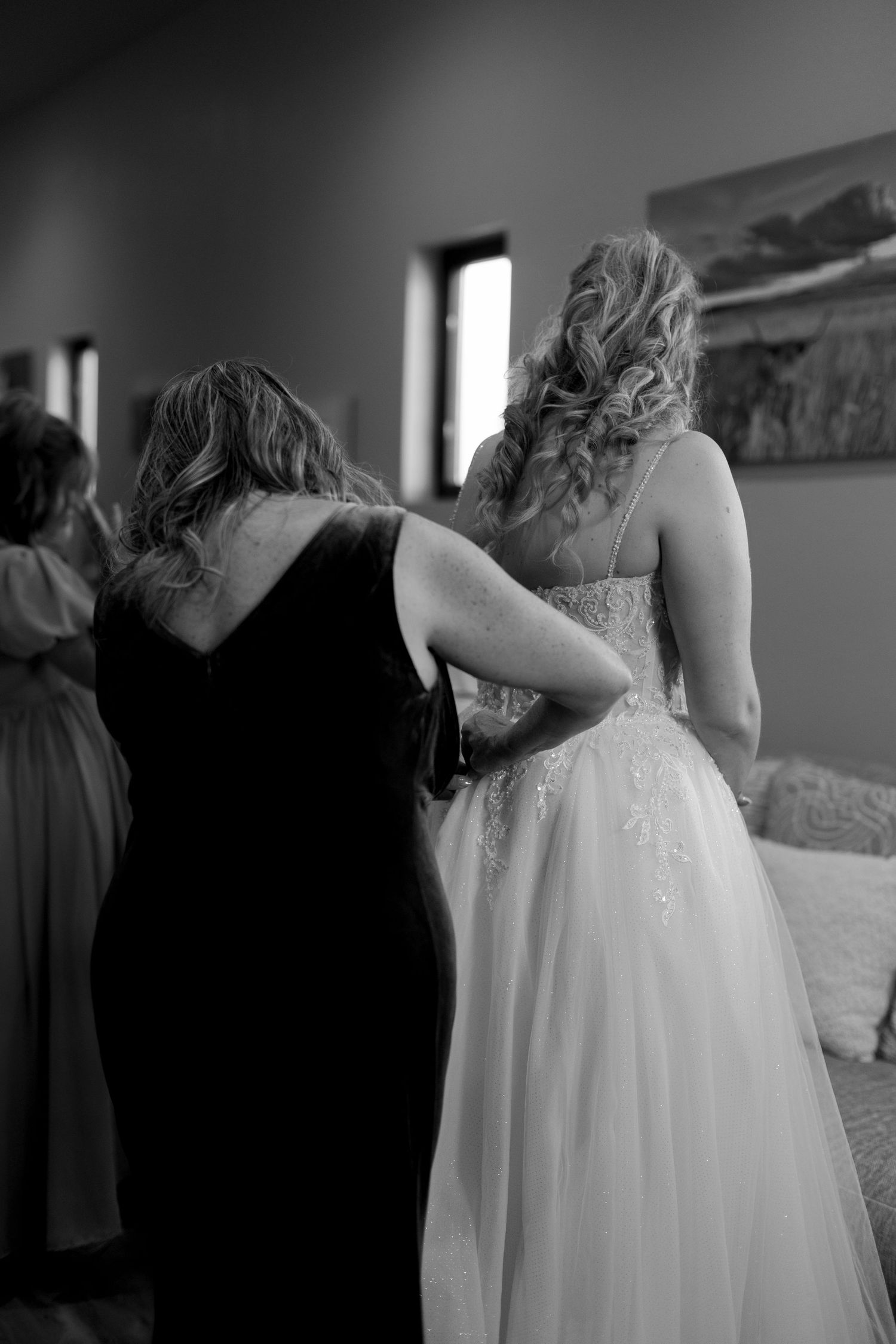 Black and white photo of bridesmaid helping bride into wedding gown before ceremony.