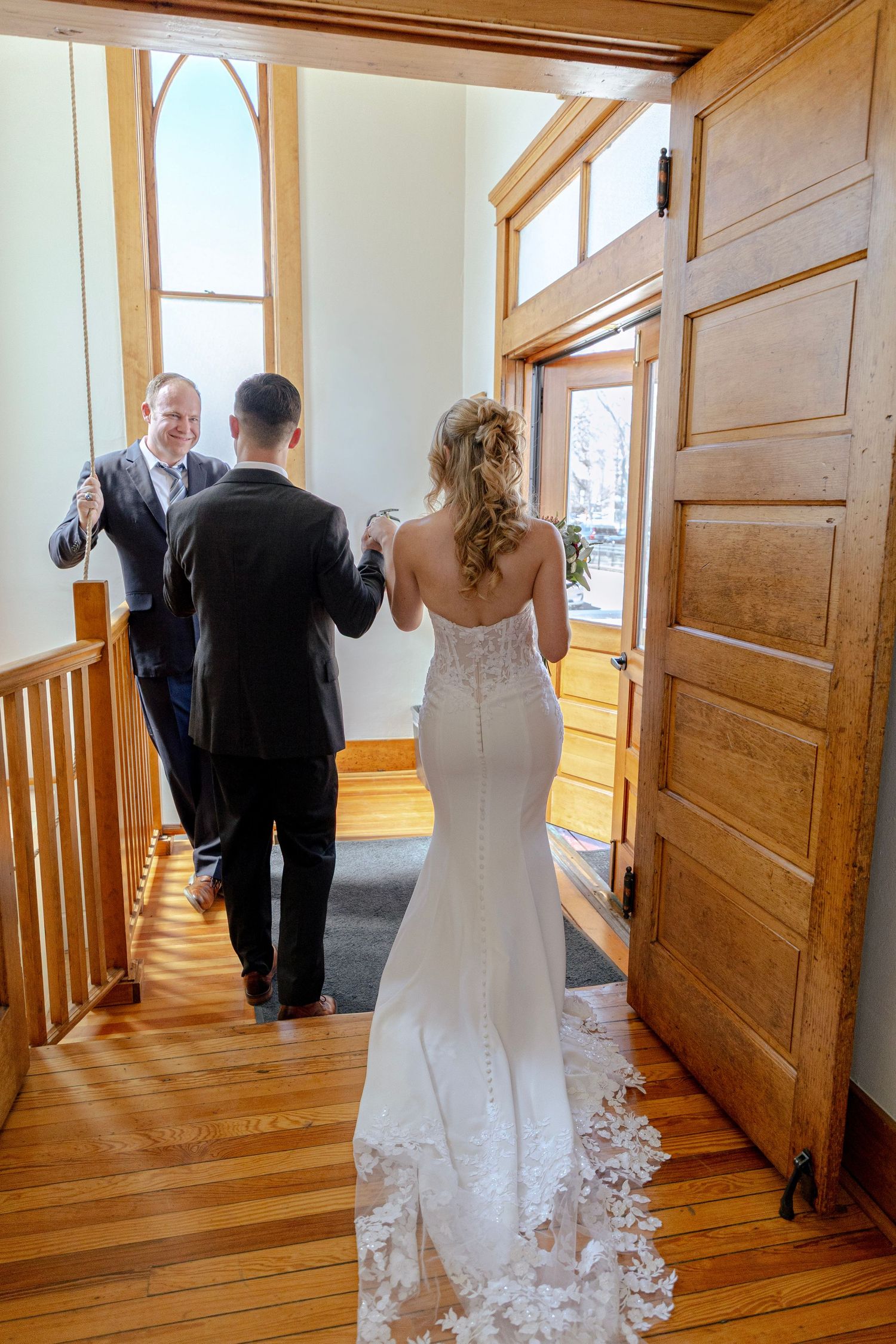 Bride in lace gown and groom enter wooden church doors as groomsman smiles in background.