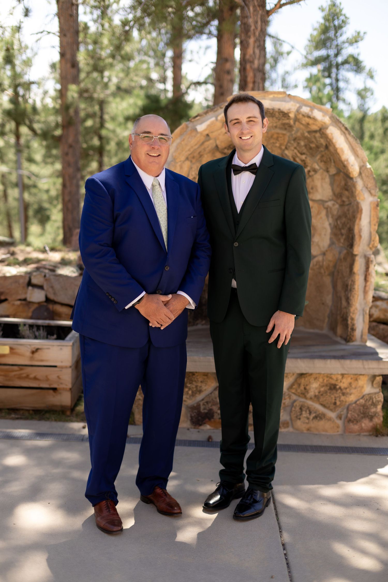Two men in formal suits, one navy blue and one dark green tuxedo, posing together at outdoor wedding venue.