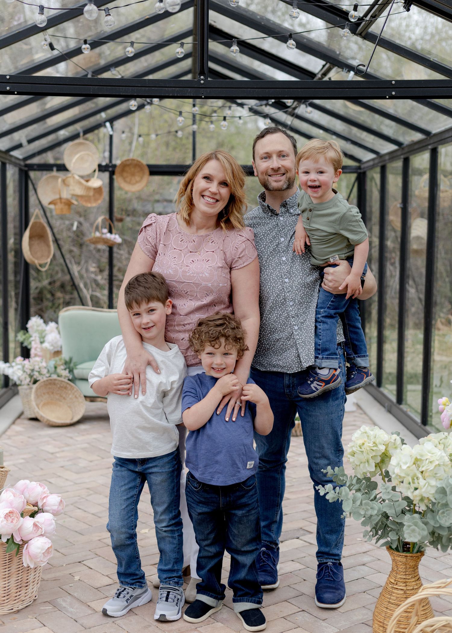 A happy family of five poses together inside a glass greenhouse with string lights and woven basket decor.