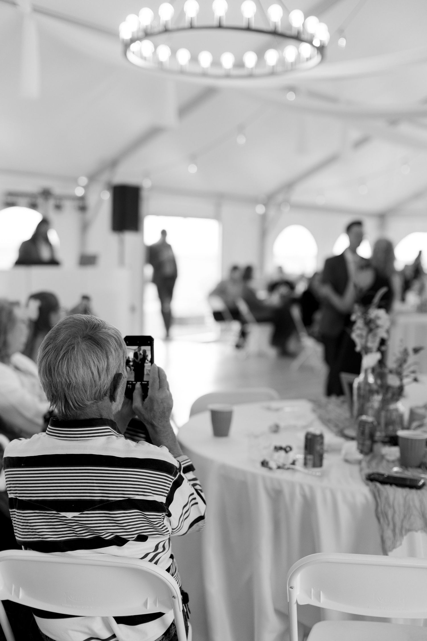 Wedding guest photographs first dance on smartphone in elegant white tent reception venue, black and white.