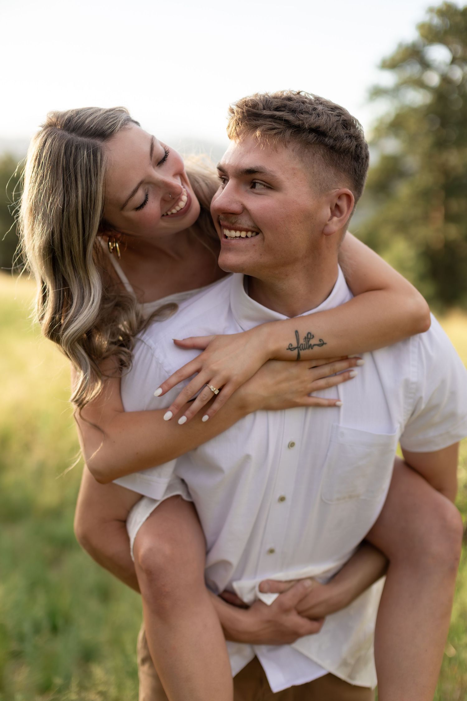 Happy couple in white outfits, man giving woman a piggyback ride in a green field, smiling and embracing outdoors.