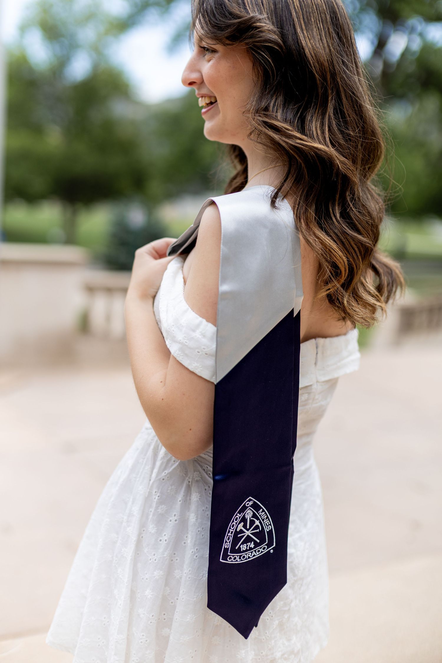 Smiling brunette woman in white dress wearing a navy and silver Colorado graduation stole outdoors.