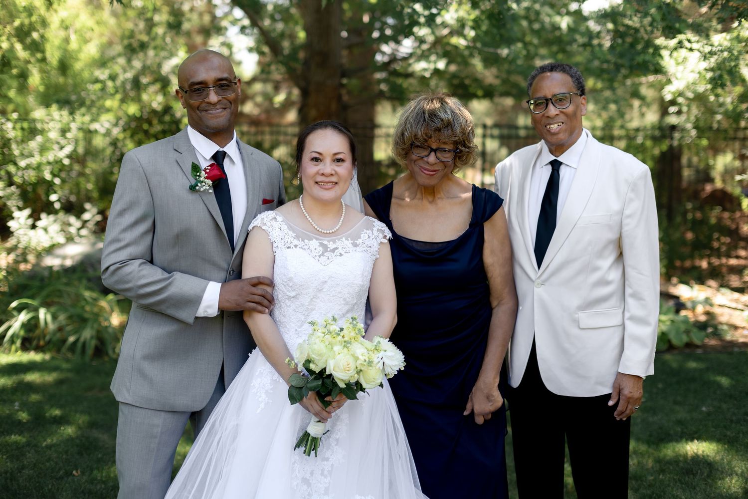A bride in a white gown holds flowers while posing with three family members outdoors at a wedding ceremony.