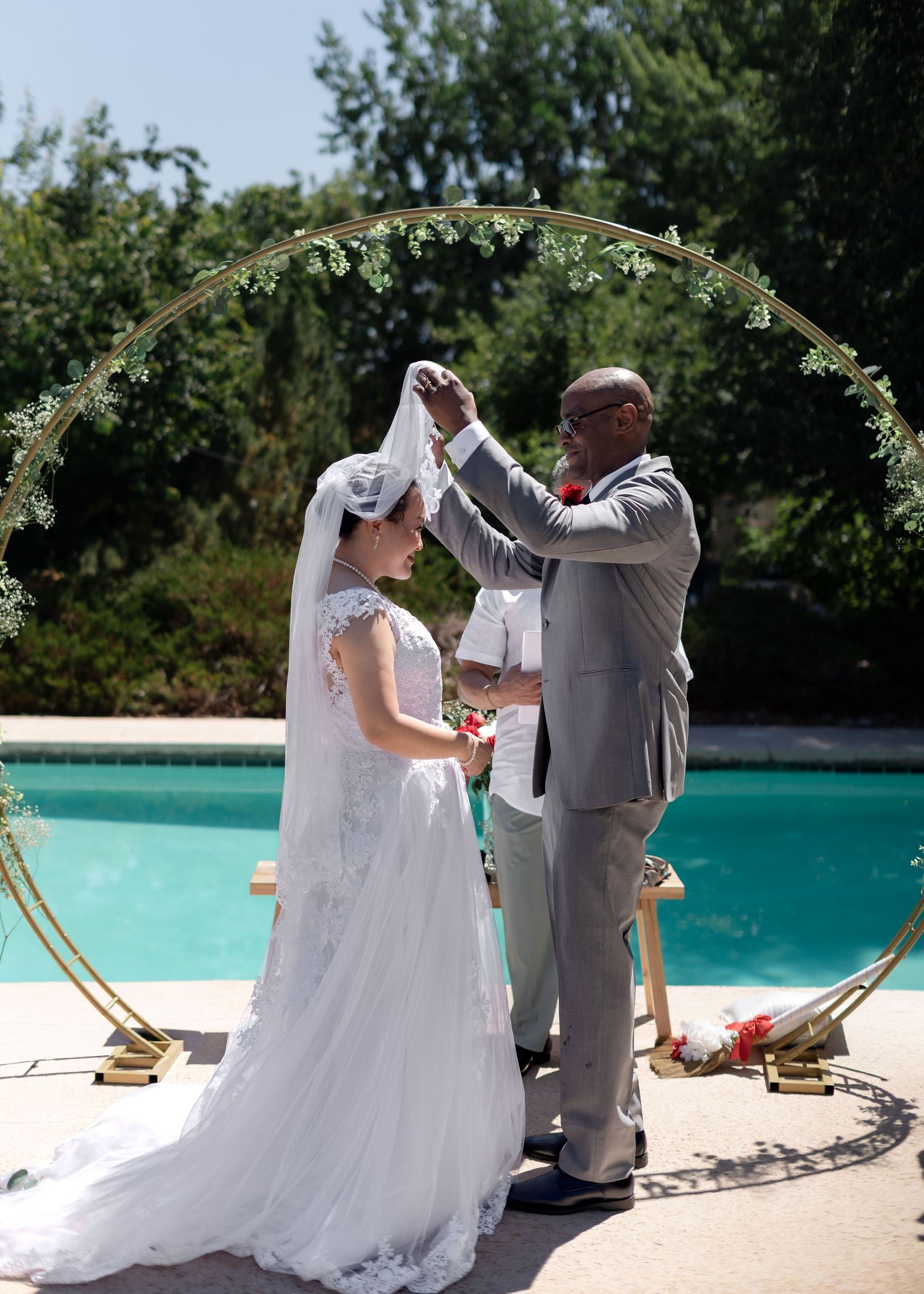 Groom lifting bride's veil during outdoor wedding ceremony by a pool under a circular floral arch.