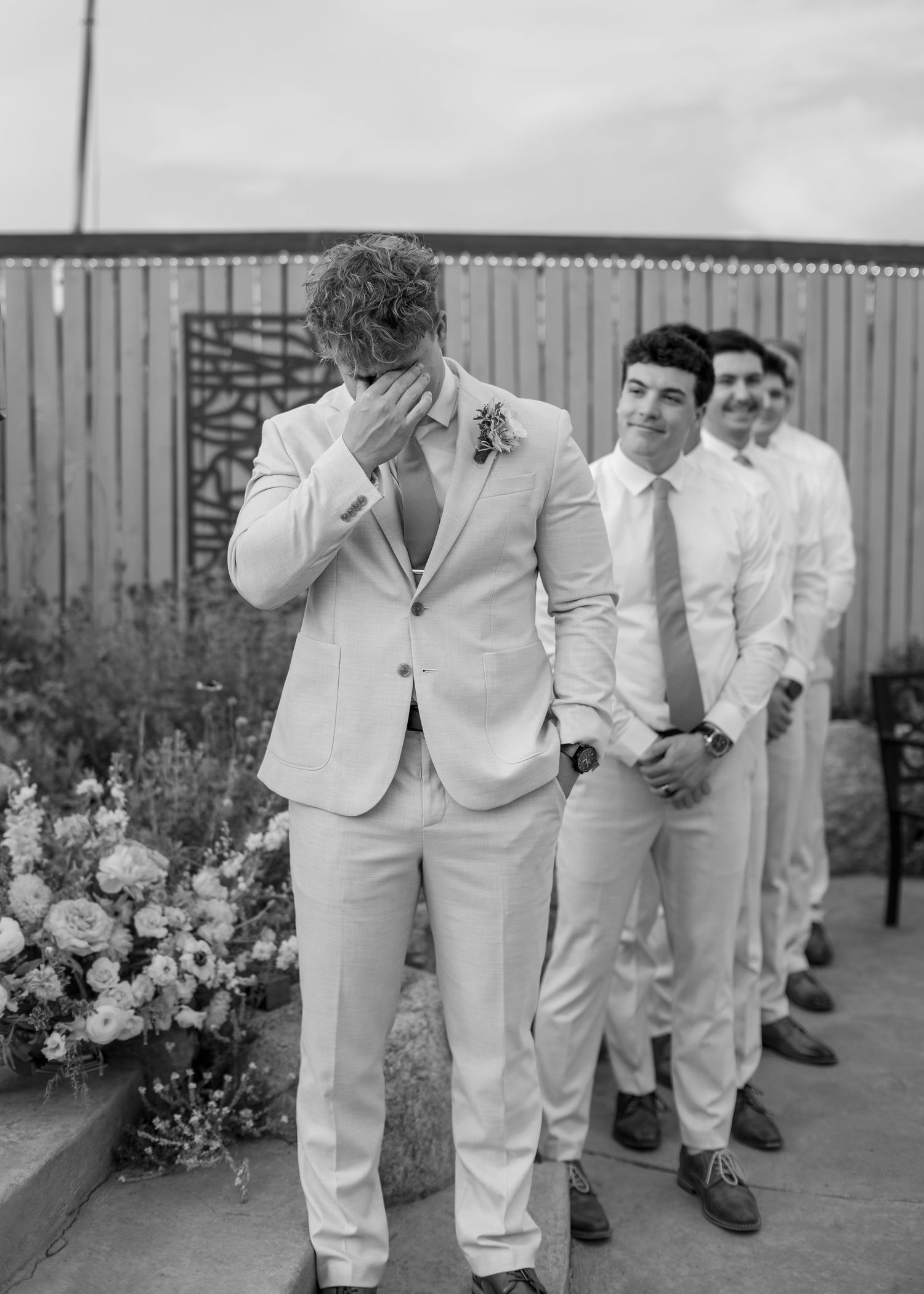 Emotional groom covers face at altar while groomsmen smile behind him in black and white photo.