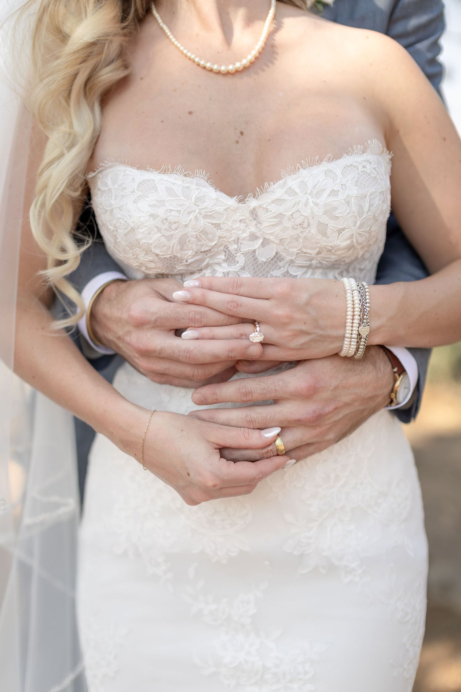 Bride and groom clasping hands at waist, showcasing diamond engagement ring, wedding bands, pearl bracelet, and white lace gown.