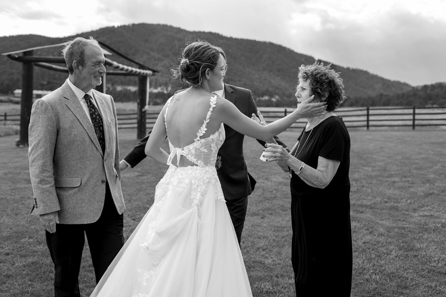 Black and white wedding photo of bride in lace gown with parents on a scenic mountain farm venue.