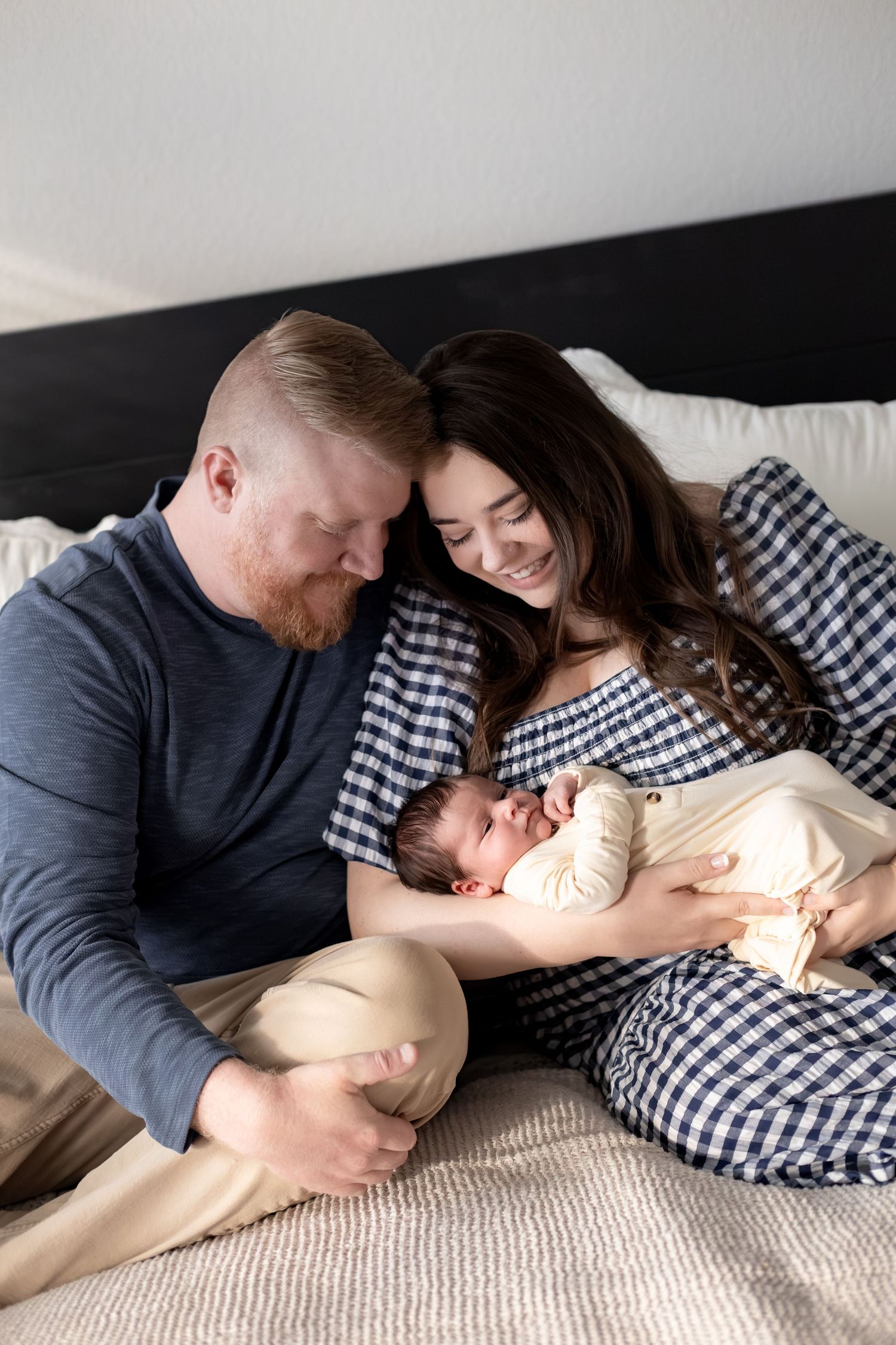 A couple smiles lovingly at their newborn baby while sitting together on a bed in a cozy bedroom setting.