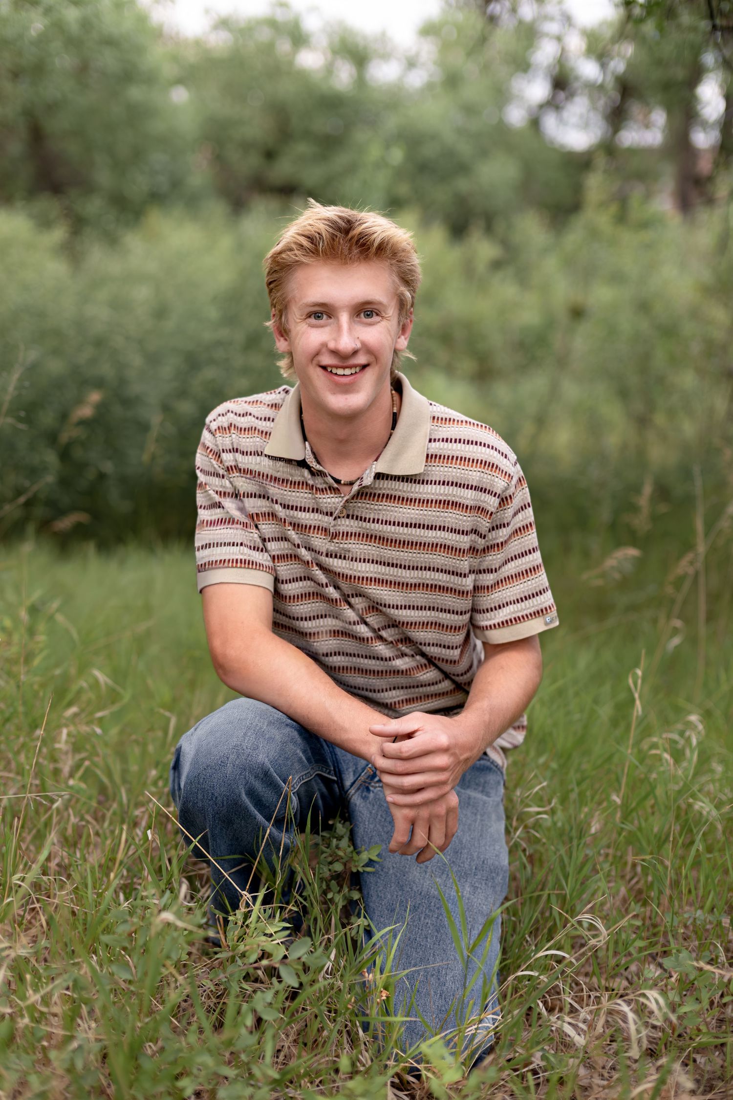 Smiling young man with blonde hair crouching in green grass wearing a striped polo shirt and jeans outdoors.