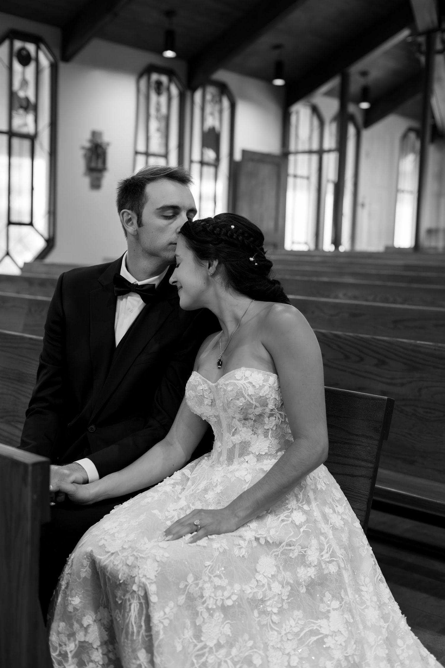 Black and white photo of a bride and groom sharing a romantic moment in a church pew, with stained glass windows behind them.