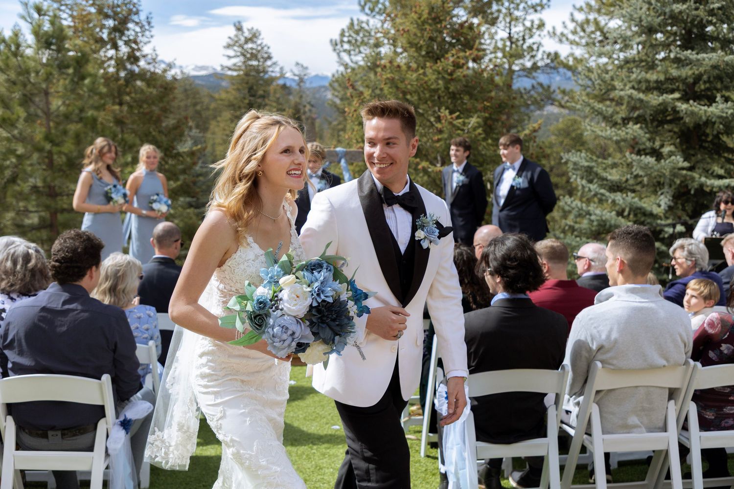 Wedding guests celebrate with dancing and laughter at an outdoor reception venue surrounded by pine trees and mountains.