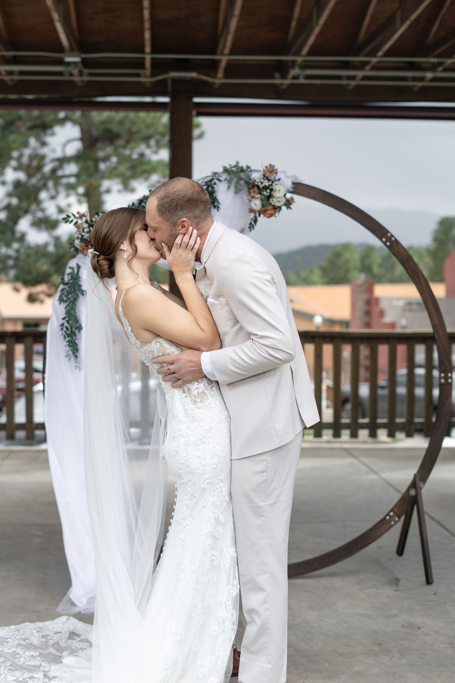 Bride and groom share first kiss at outdoor wedding ceremony beneath circular floral arch with mountain views.