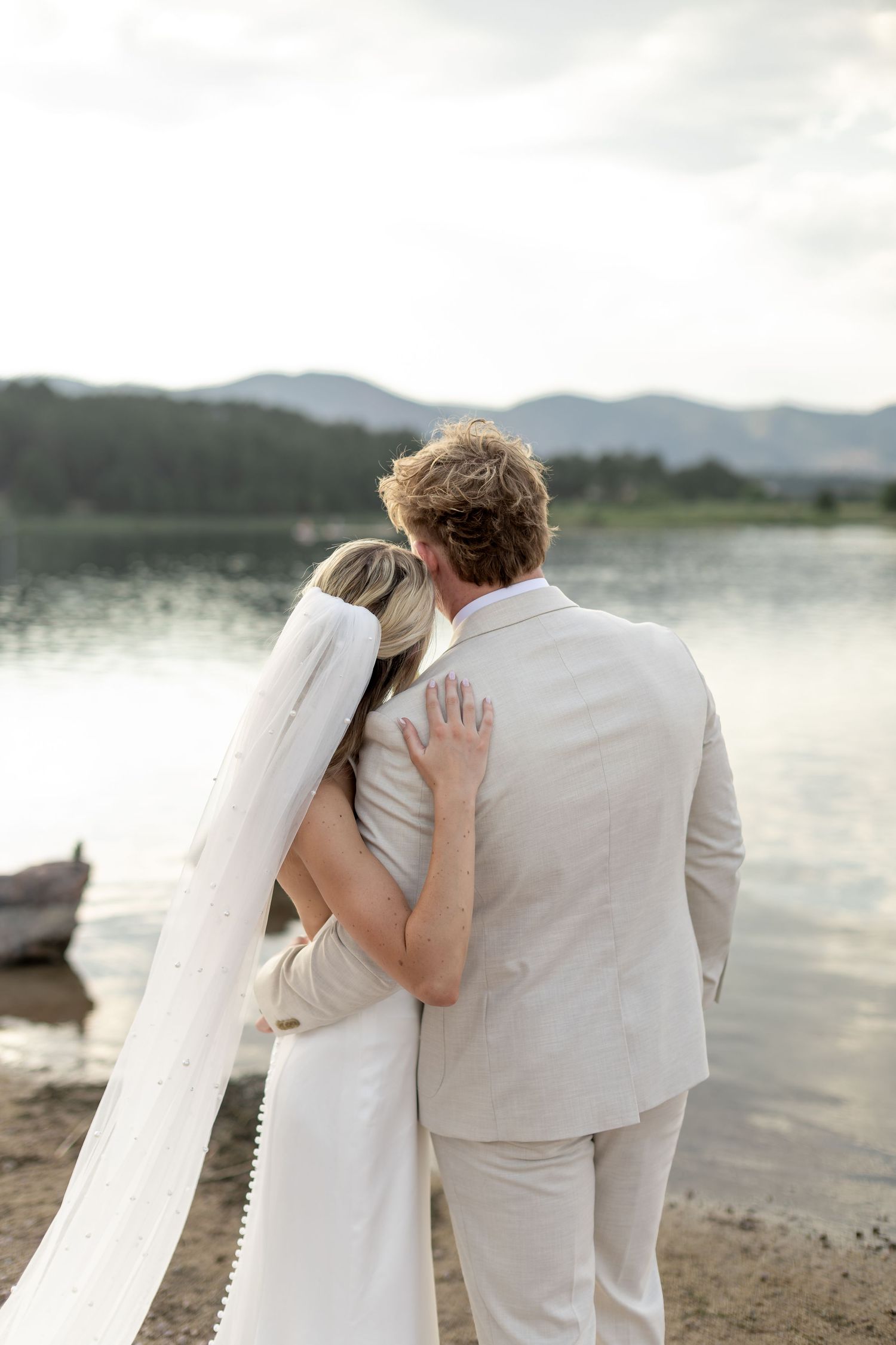 Bride and groom embrace facing a serene lake with mountain views on their wedding day.